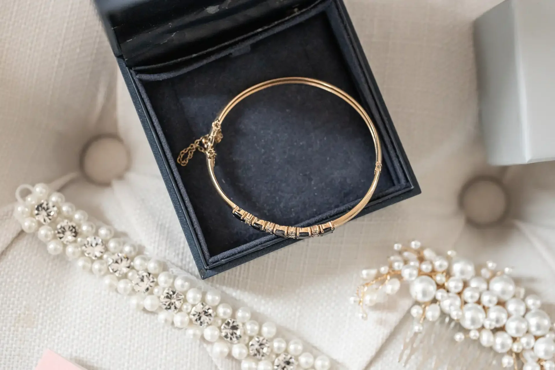 A gold bracelet in a black box on a cushioned surface, surrounded by a pearl bracelet and pearl earrings.