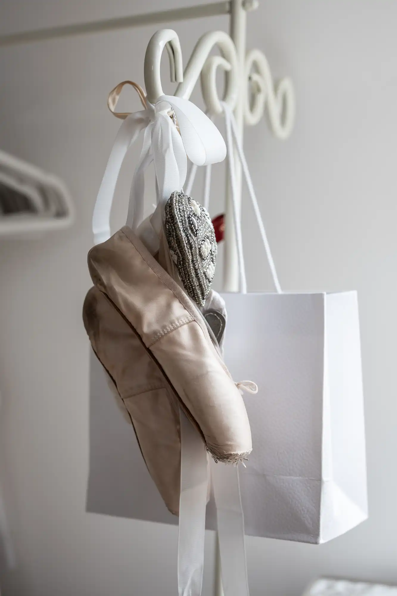 Ballet pointe shoes with ribbons and a decorative item hang on a white stand next to a paper bag in a softly lit room.