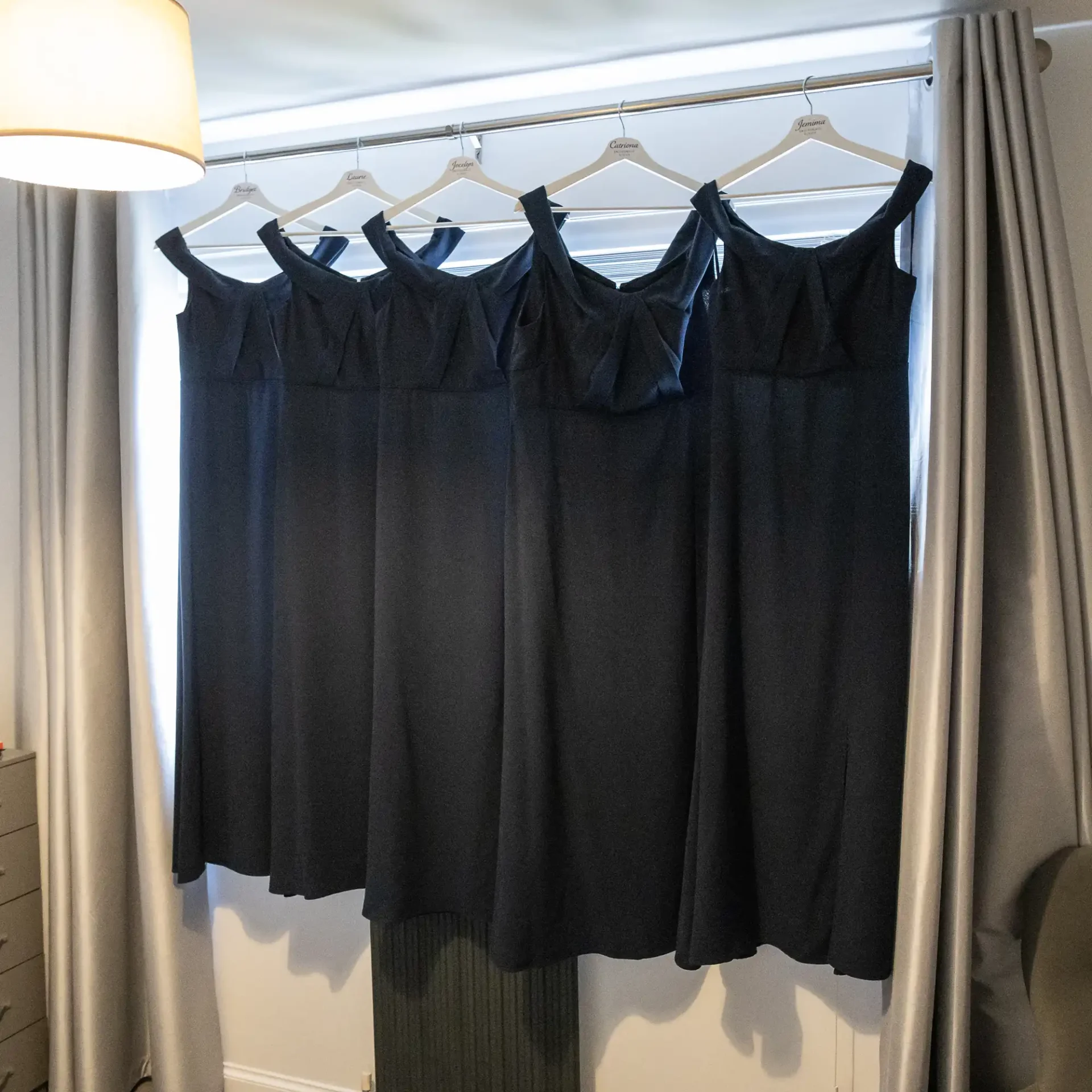 Five identical navy blue dresses hang on hangers on a silver rod in front of a window with light gray curtains.