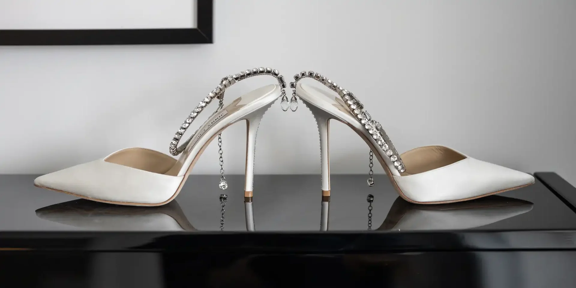 White high-heel shoes with crystal-embellished straps displayed on a glossy black surface against a light gray background.