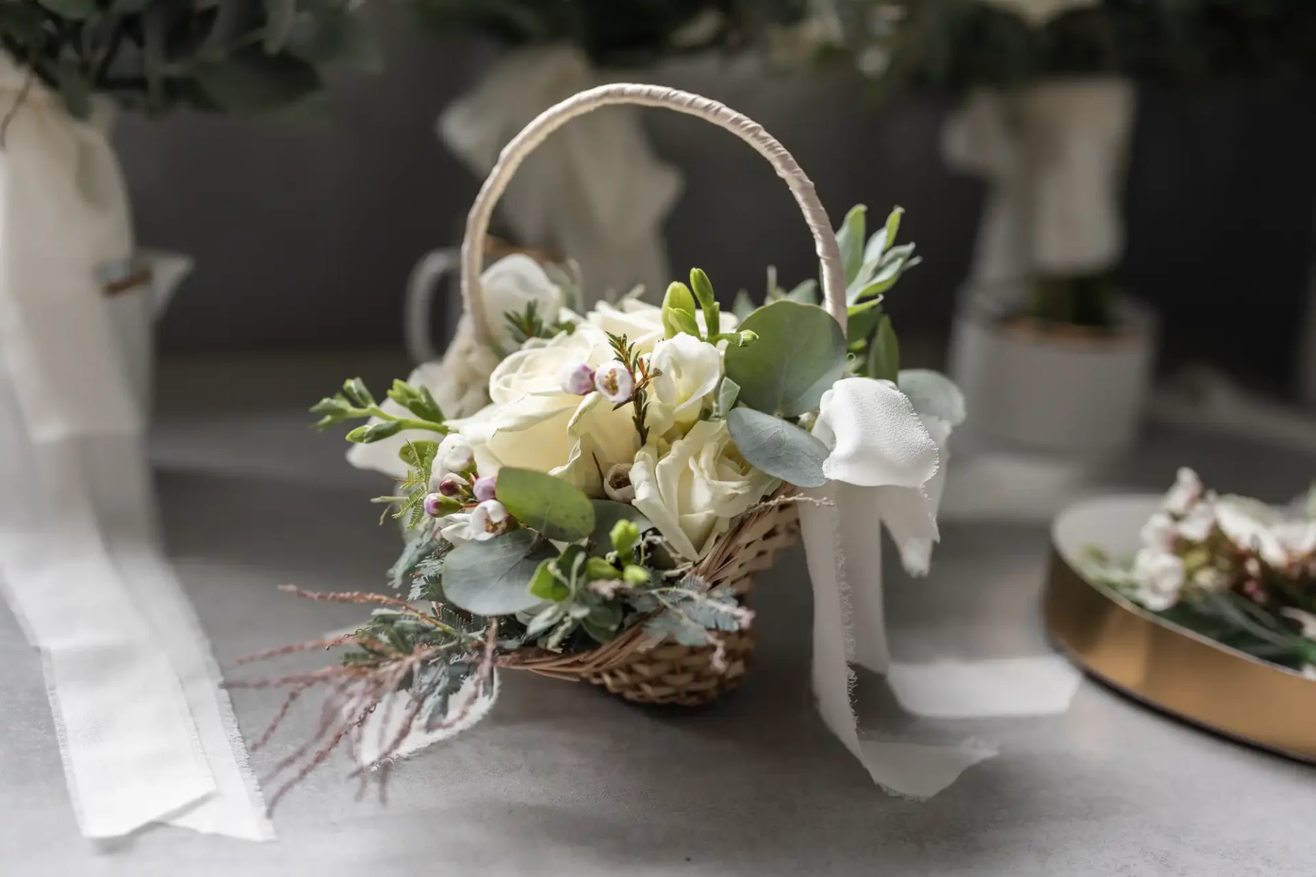 A small woven basket filled with white roses, eucalyptus, and greenery, adorned with a white ribbon, placed on a gray surface.