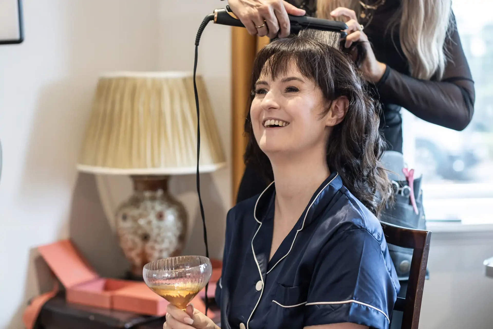 A woman in a navy blue pajama set holds a glass while having her hair styled with a straightener. A decorative lamp and orange boxes are in the background.