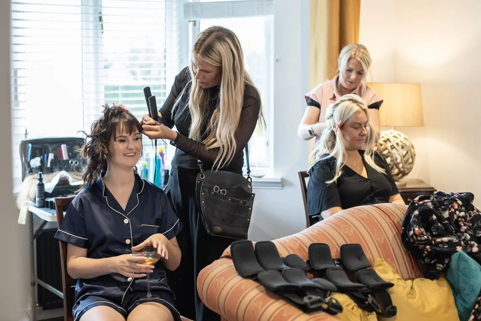 Two women sit in a room getting their hair styled by two hairstylists. One holding a drink, the other with hair in curlers. A hairdressing tool set is on a table nearby.