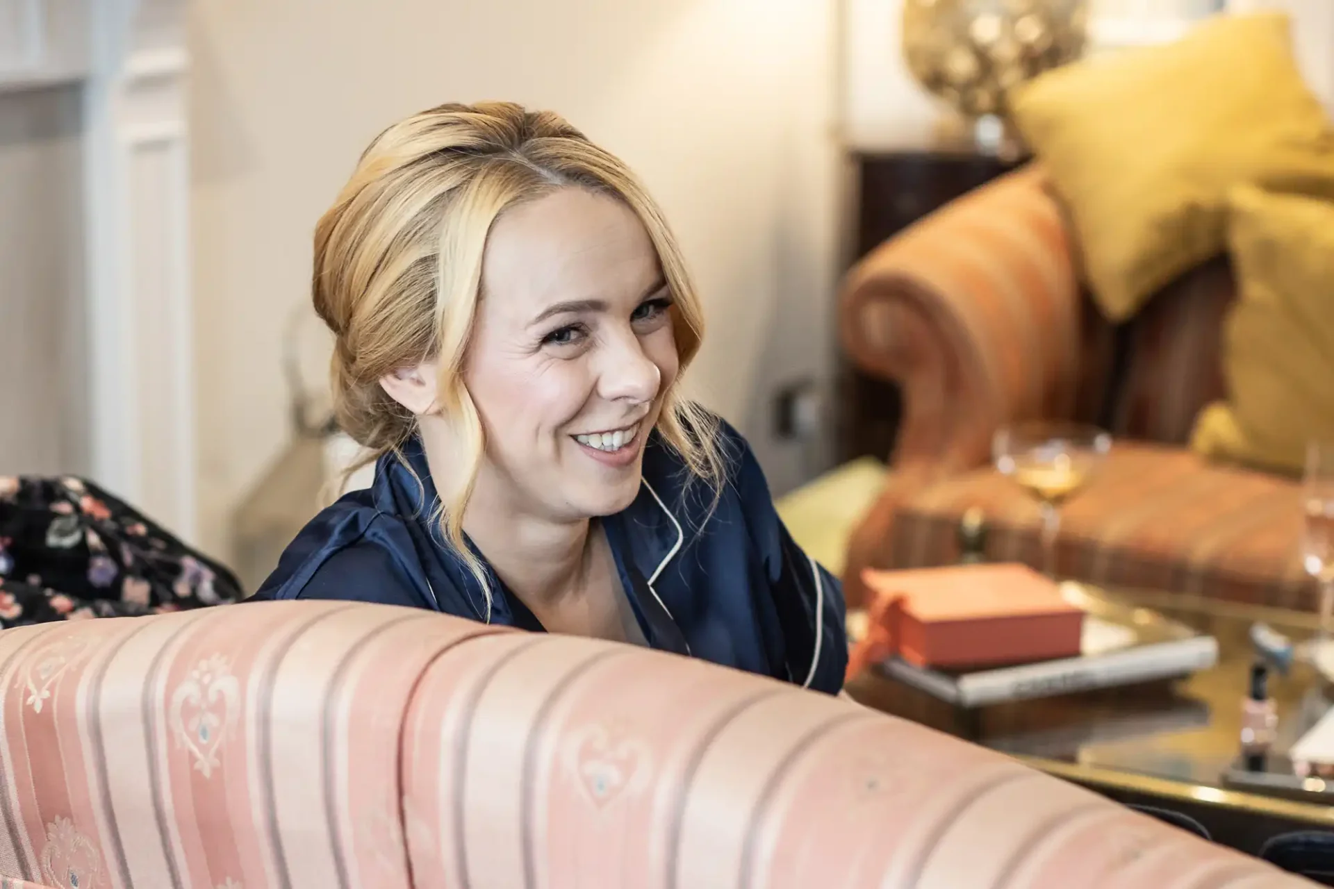 A woman with blonde hair smiles while sitting on a pink patterned sofa. There are cushions and a glass table with books and a cocktail glass in the background.