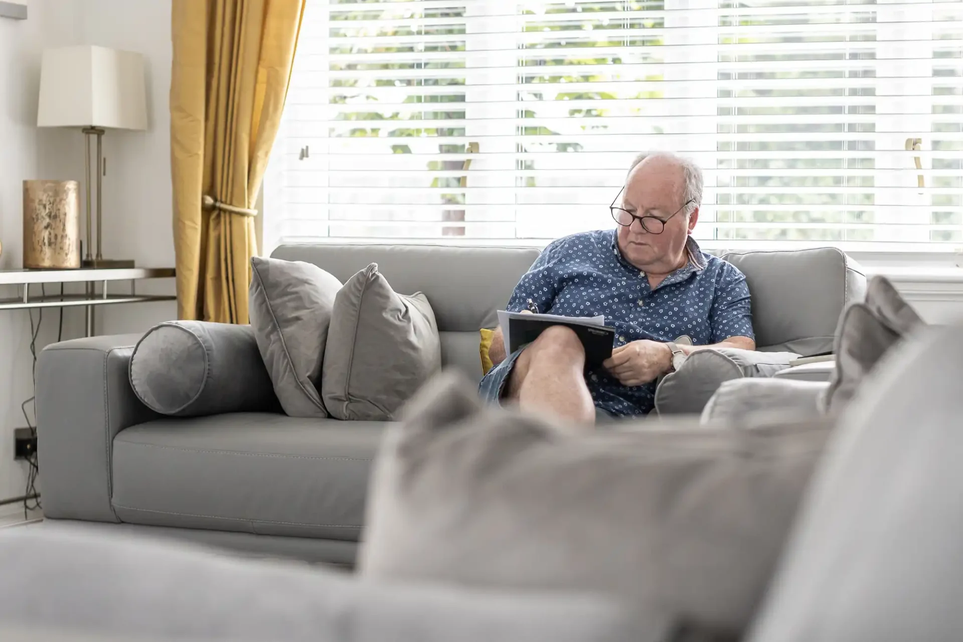 Man sitting on a gray sofa in a living room, reading a tablet. Large windows with blinds and yellow curtains are in the background.