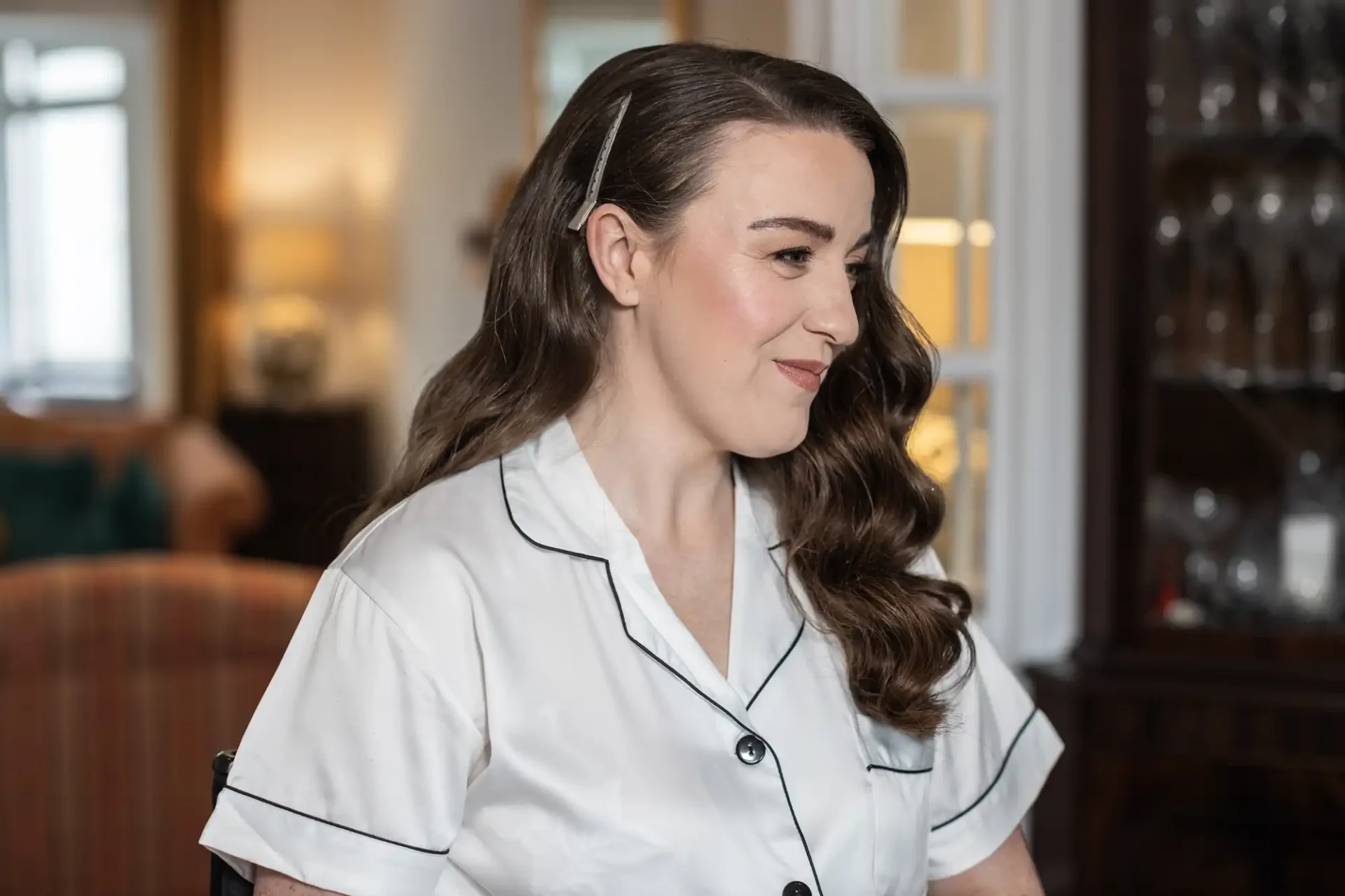 Woman with long wavy hair wearing a white collared shirt, sitting indoors near a cabinet, smiling softly.