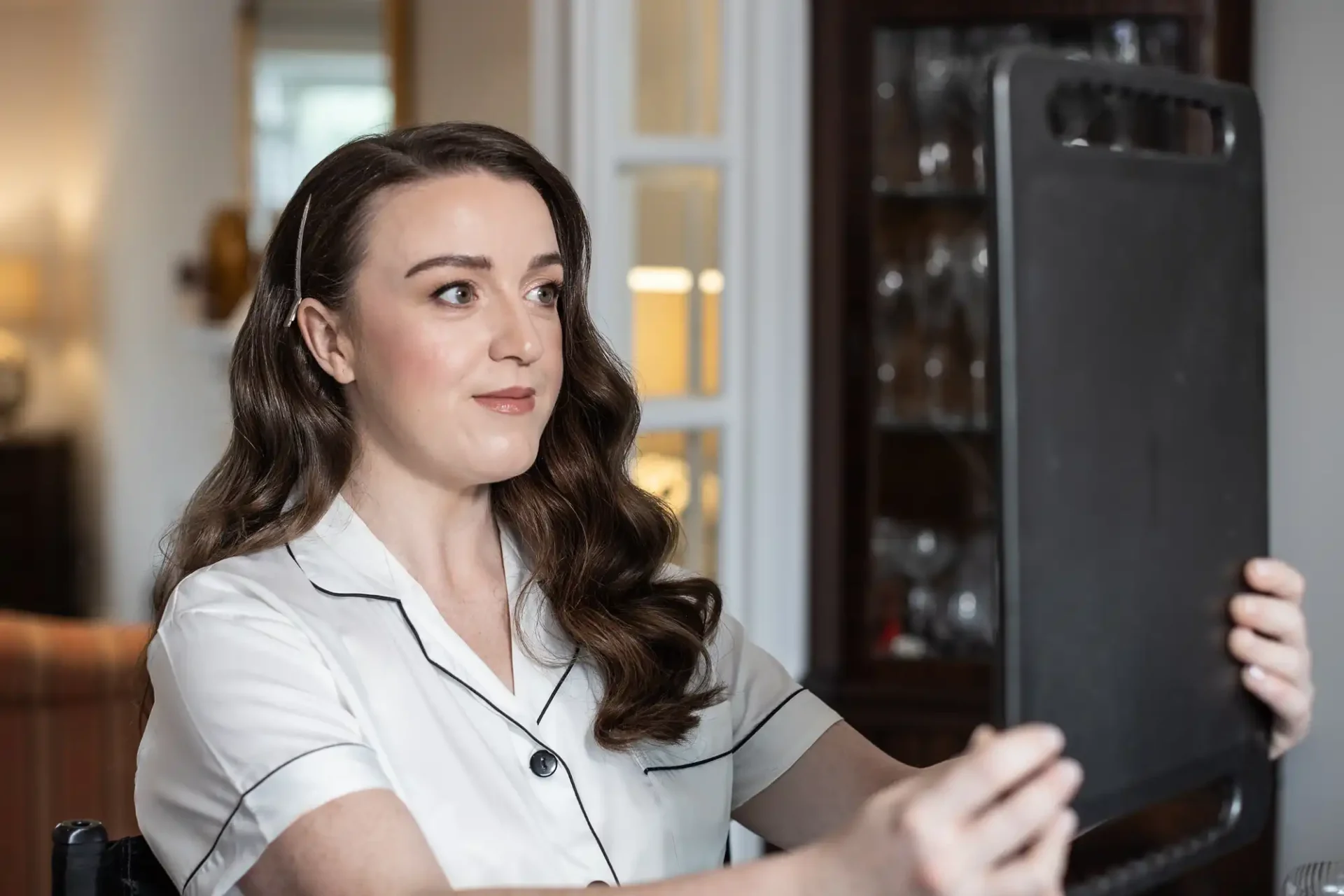 A woman with long brown hair in a white shirt sitting indoors, holding and looking at a large rectangular mirror.