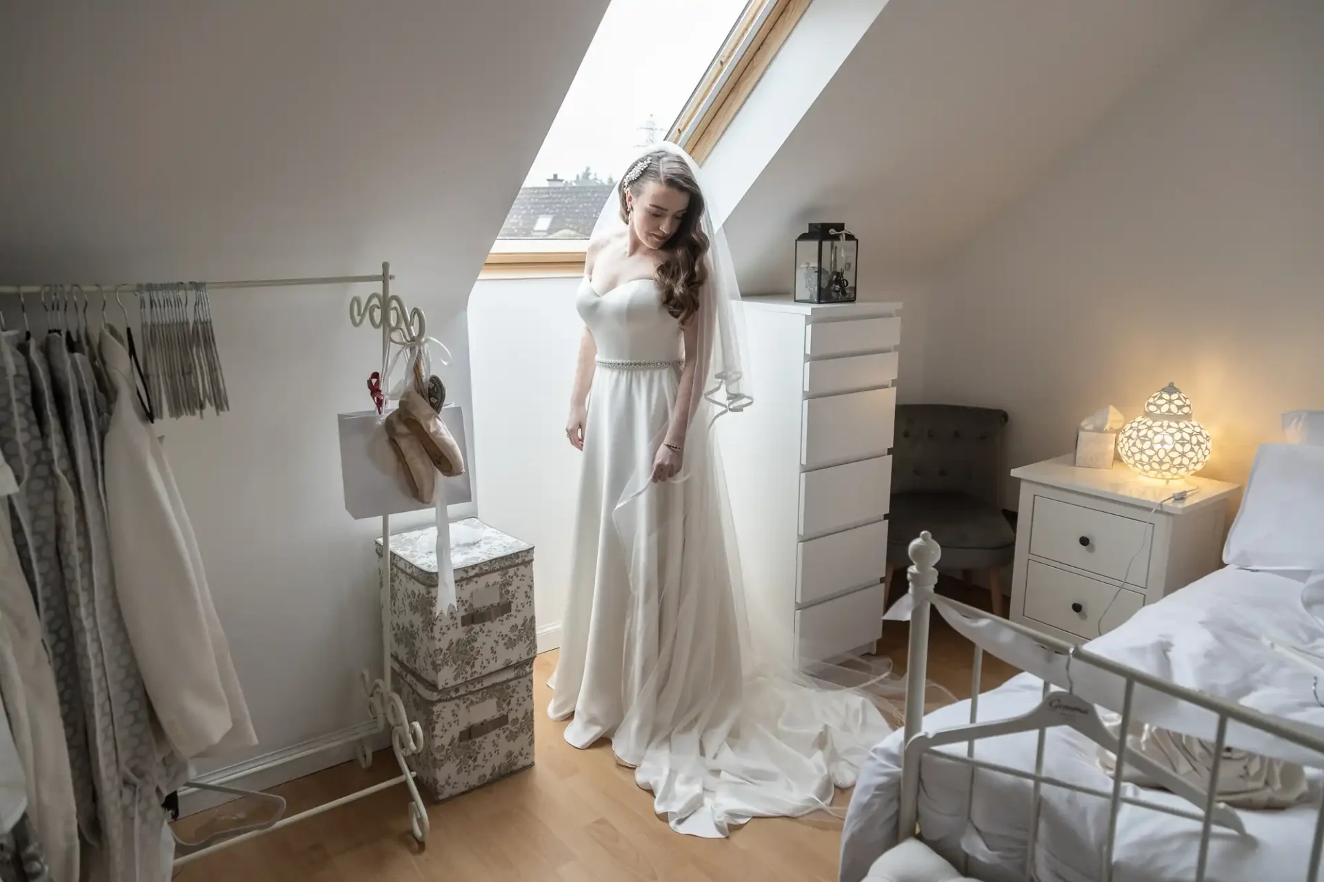 Bride in a white wedding dress stands near a window in a bright attic room with a white dresser and a bed.