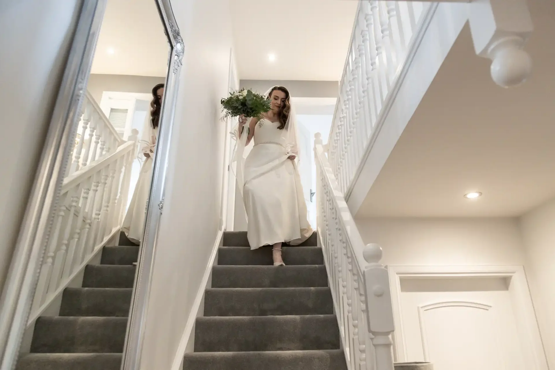 A bride in a white dress descends carpeted stairs holding a bouquet, with white railings and walls surrounding her.