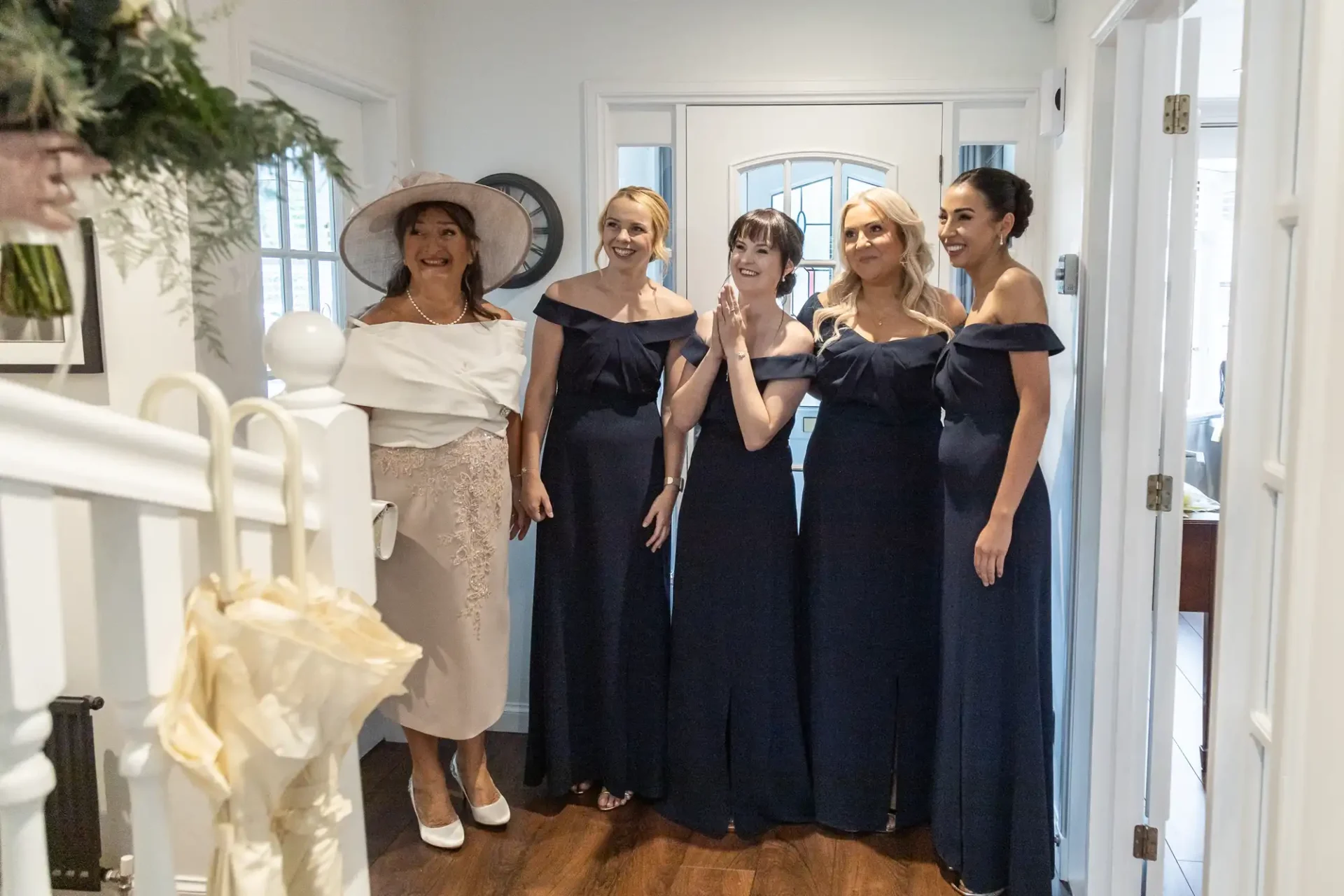 Five women in formal attire stand indoors, four in navy dresses and one in a light, stylish outfit with a hat, smiling and looking towards a staircase.