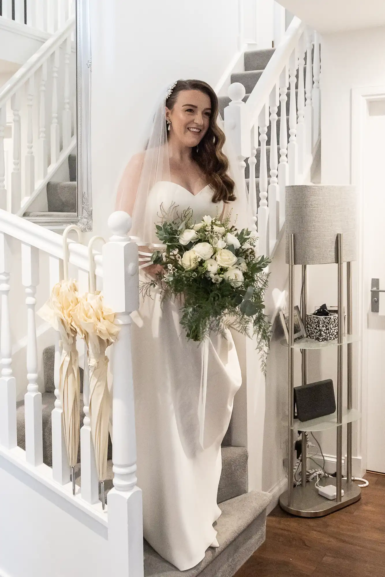 A bride in a white gown holds a bouquet of white roses while descending a staircase decorated with flowers.