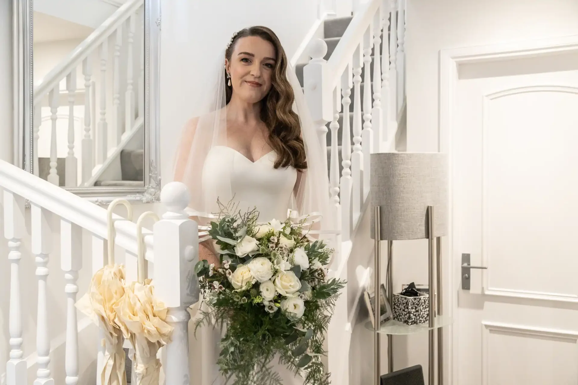 A bride in a white dress stands on a staircase, holding a bouquet of white and green flowers. She smiles, with a lamp and mirror in the background.