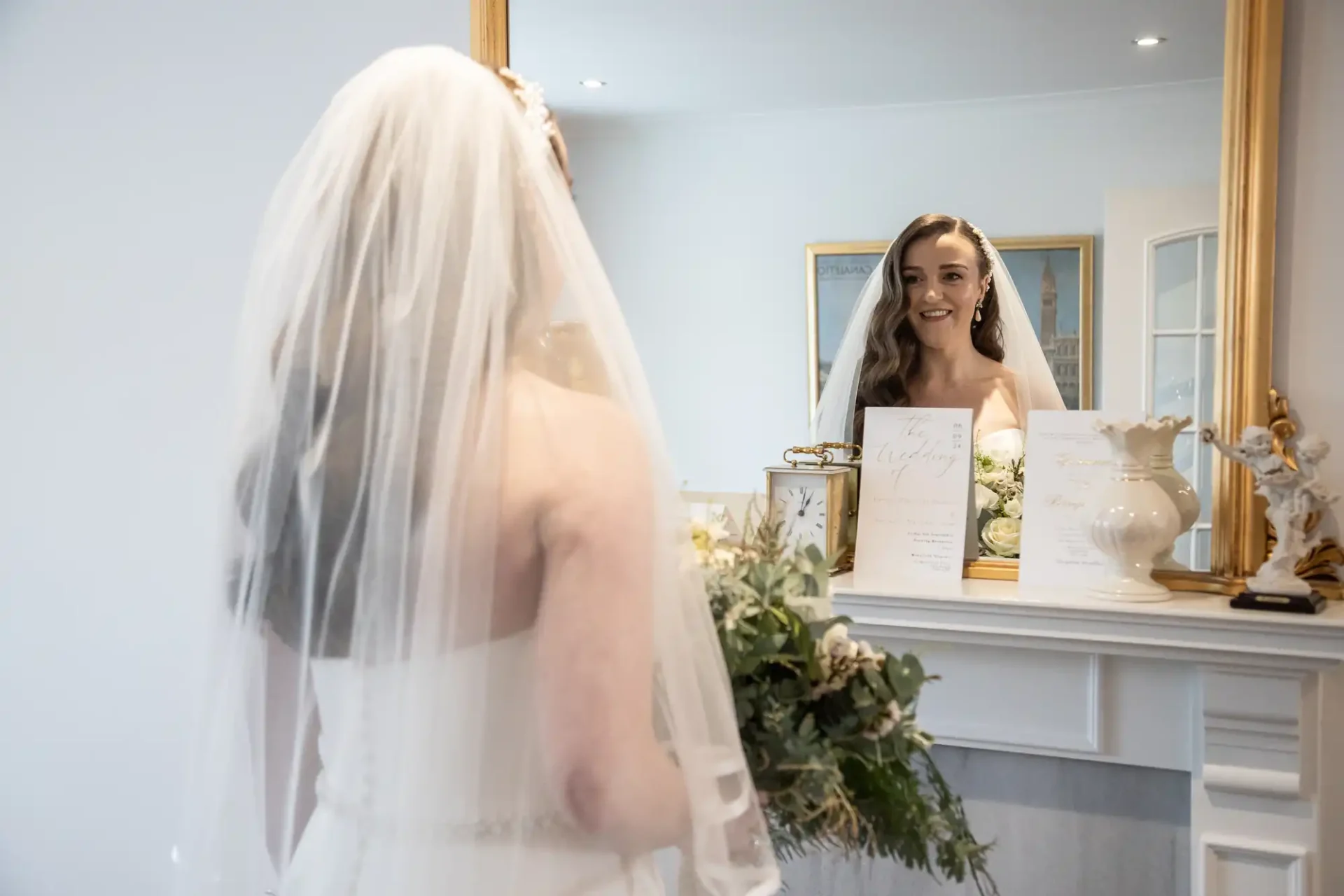 Bride in white gown and veil holding a bouquet, looks at her reflection in a mirror above a mantelpiece, with wedding invitations and decor on the mantel.