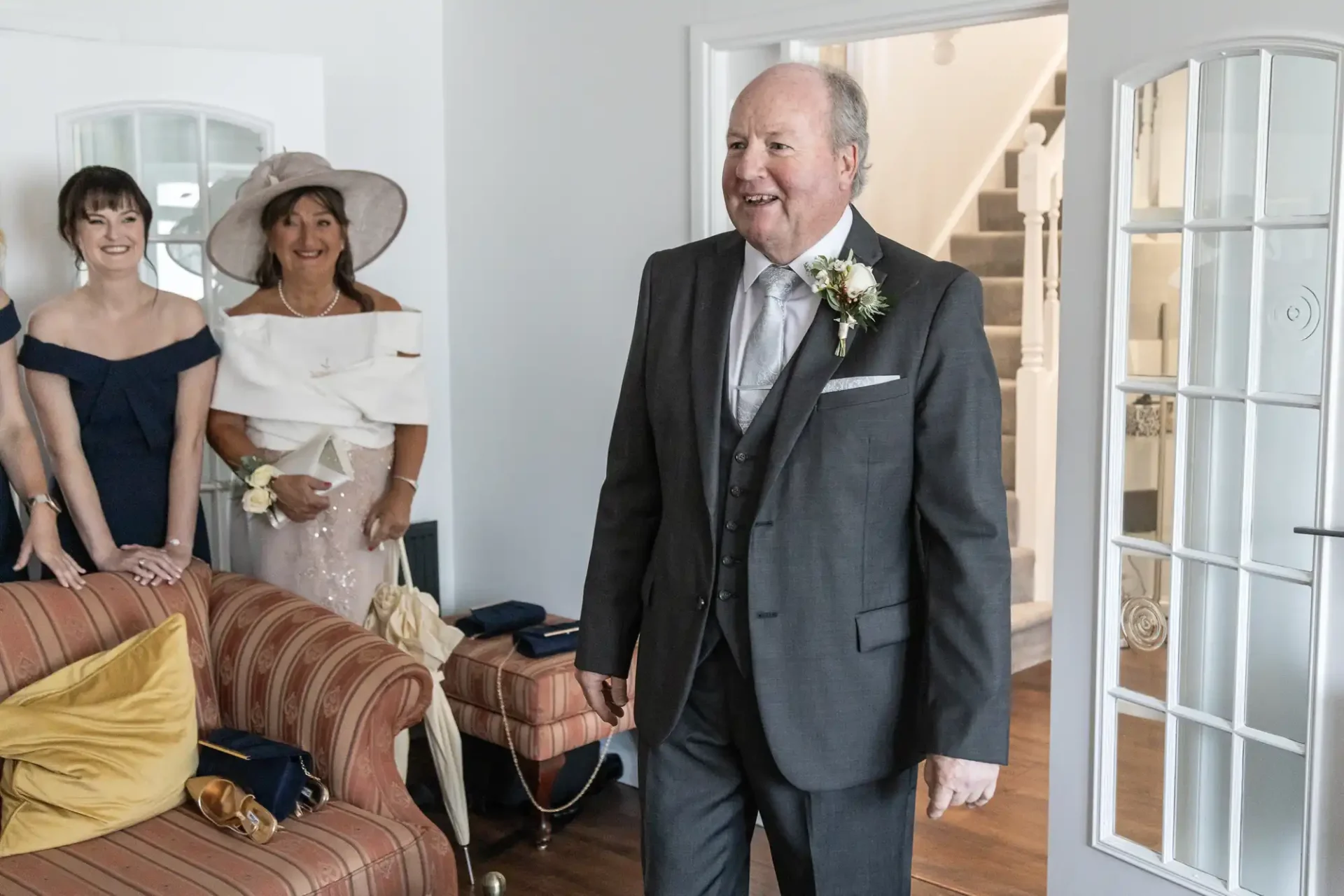 A man in a suit and tie, with a boutonniere, stands smiling in a living room. Two women, one in a hat and the other in a dress, watch from a couch.