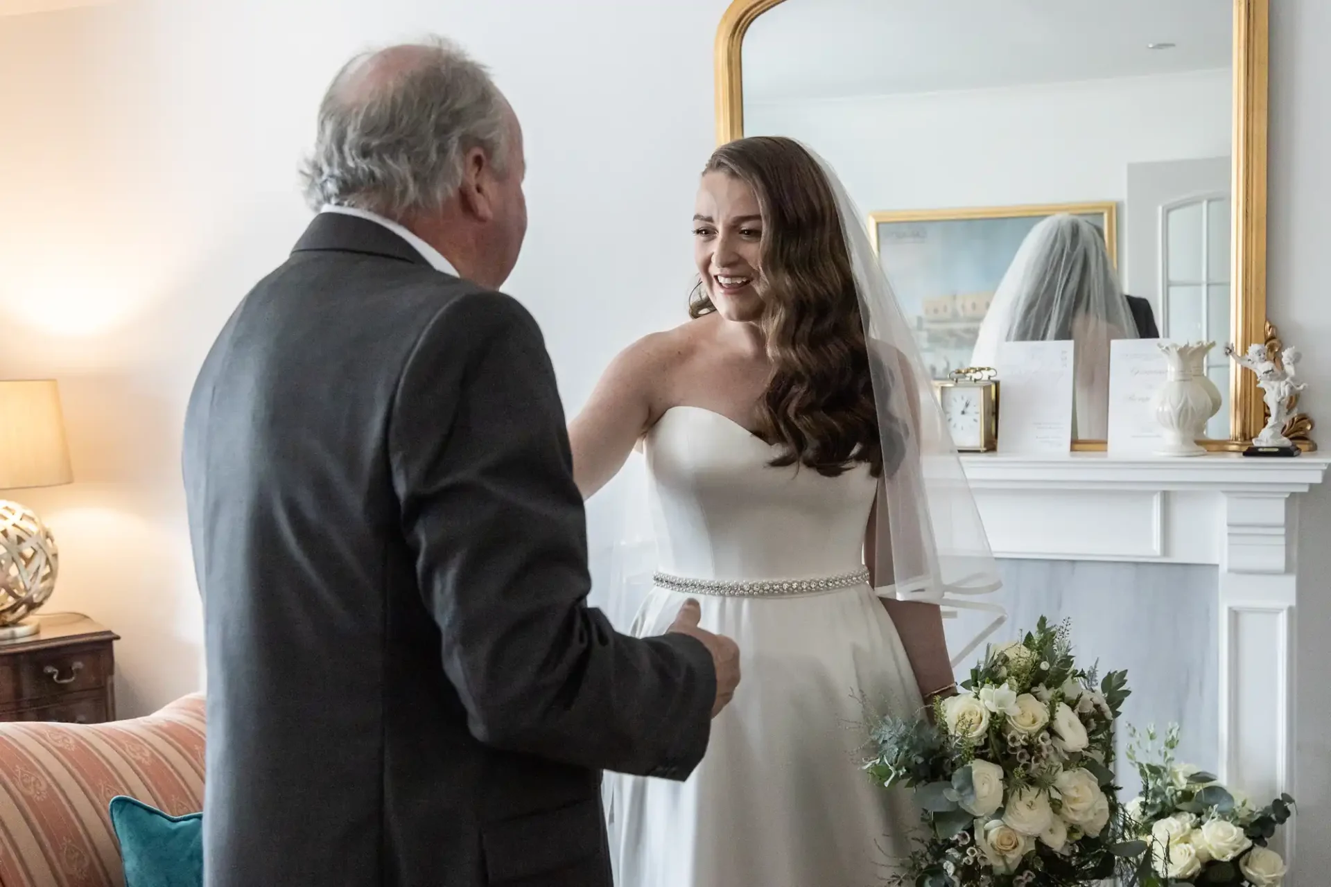 A bride in a white dress stands next to an older man in a suit in a living room, smiling. Flowers are on a nearby table.