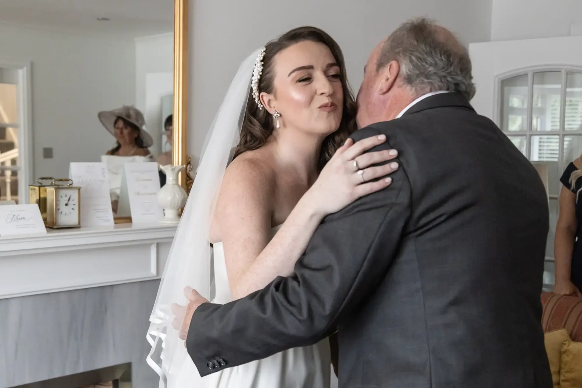 Bride in a white dress and veil embraces an older man in a suit in a living room setting. A large mirror and decorative items are visible in the background.