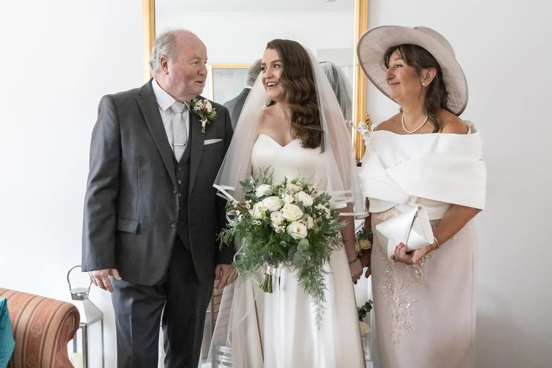 Bride in a white gown holding a bouquet, stands between a man in a suit and a woman in a dress and hat, in front of a mirror.