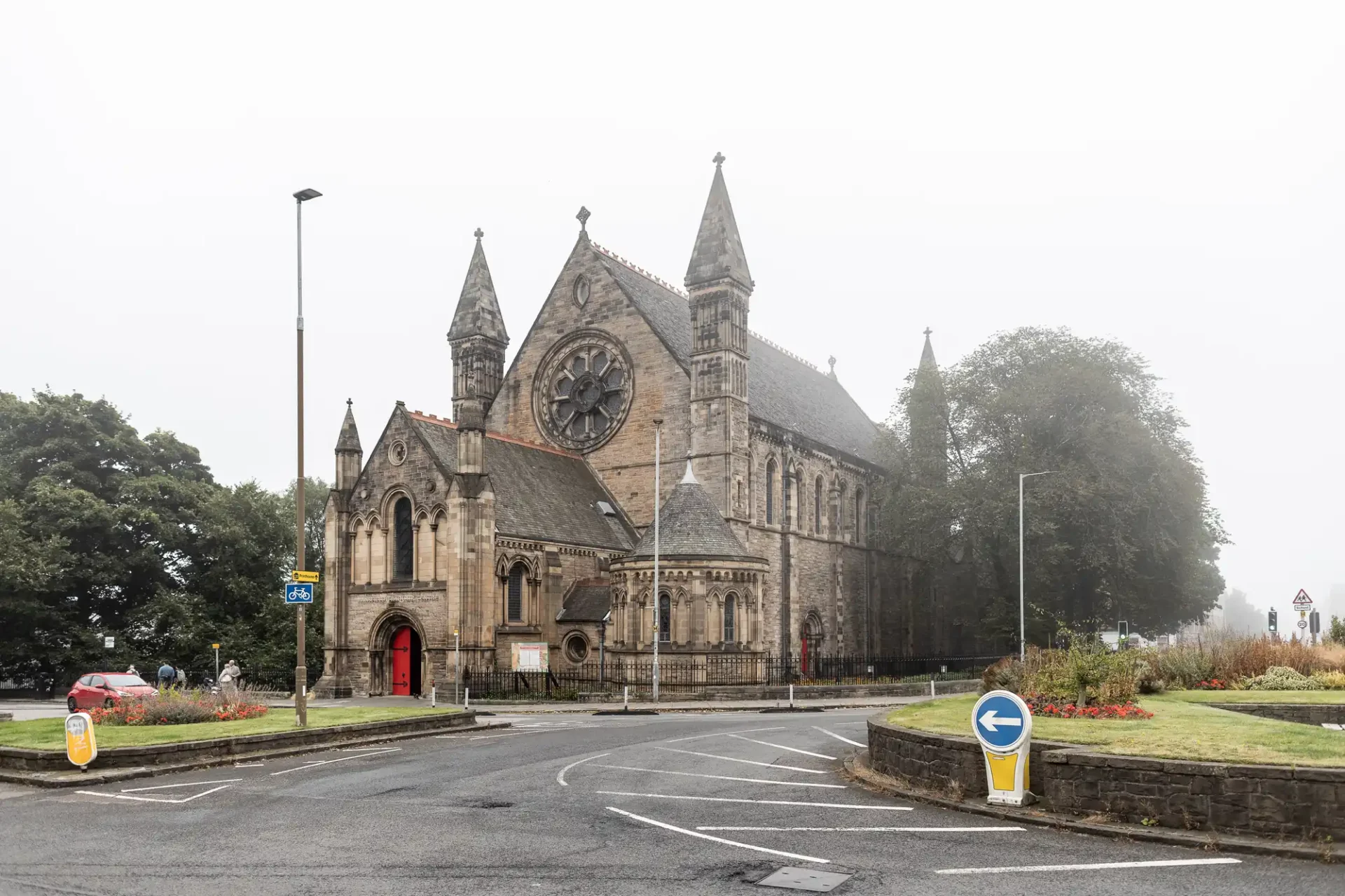 Gothic-style church with a large rose window, surrounded by trees and fog. Road signs and a small roundabout are in the foreground.