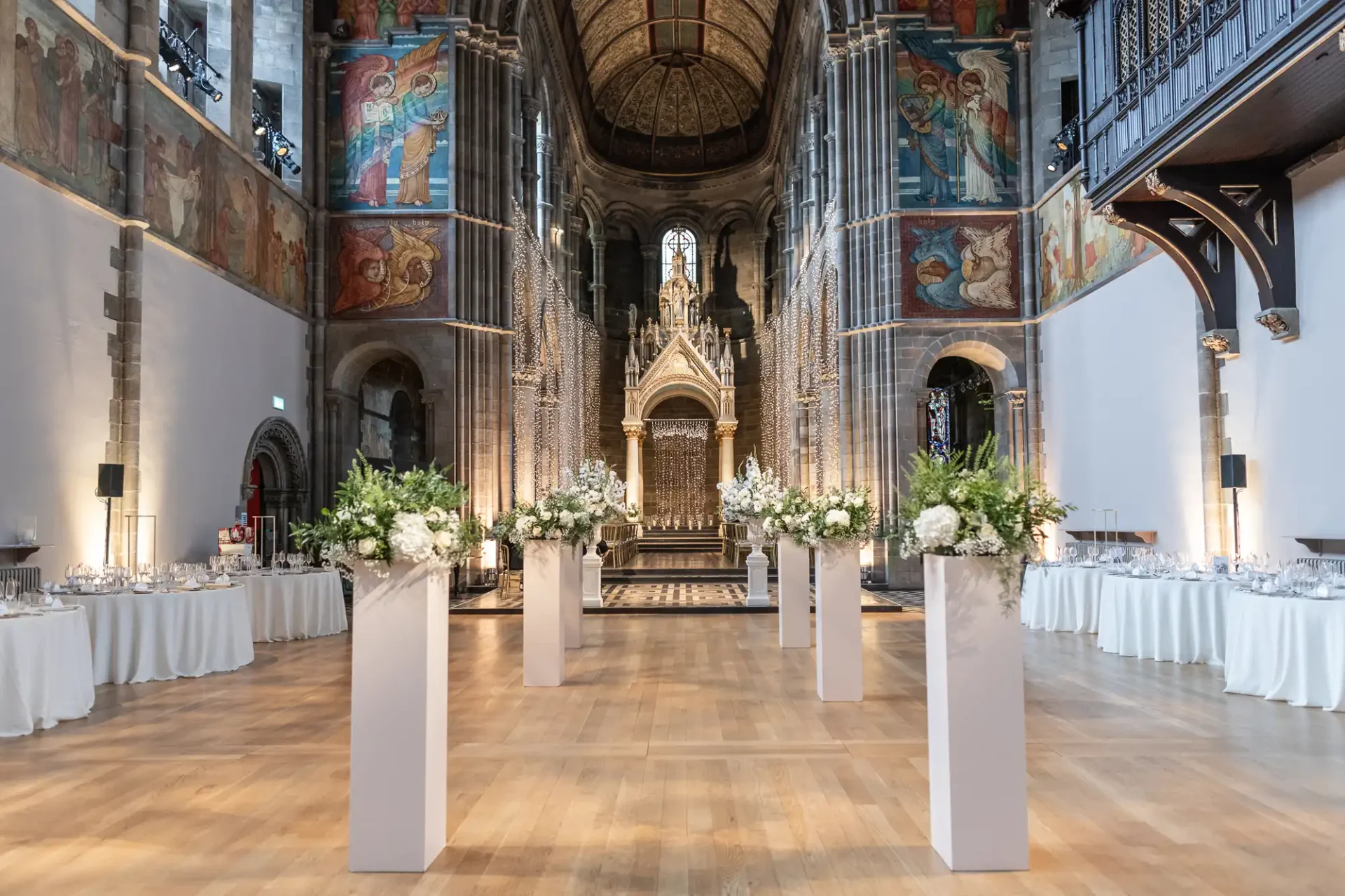 A grand hall with ornate architecture, decorated with tall flower arrangements and set tables, likely prepared for a formal event or ceremony.