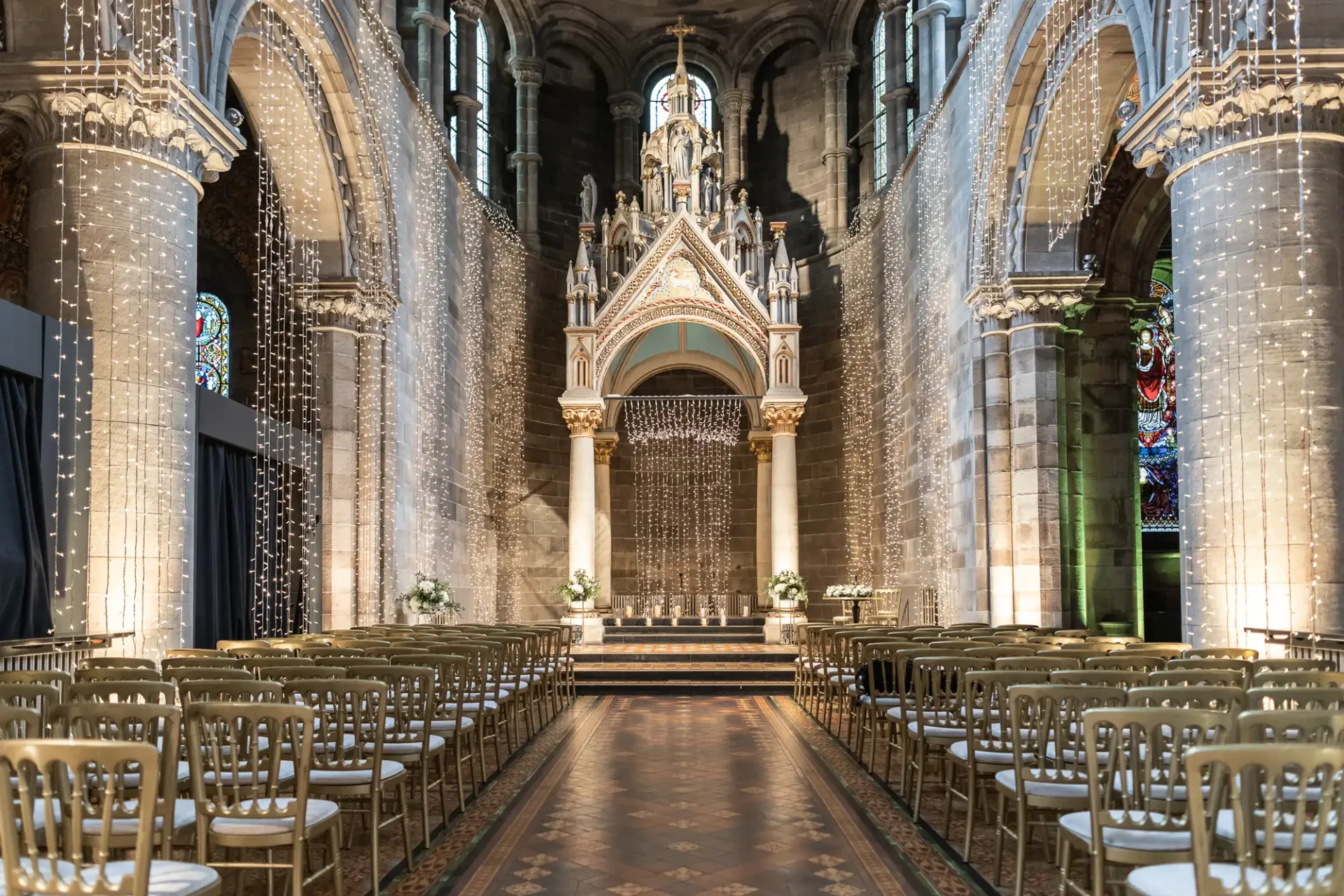 Ornate church interior set for a wedding, with rows of gold chairs, decorative arches, stained glass windows, and string lights.