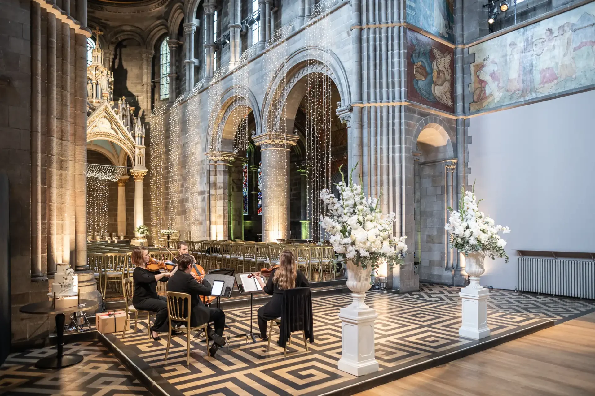 Musicians perform in a historic church interior with stone arches and ornate decor. White floral arrangements on pedestals flank the scene.