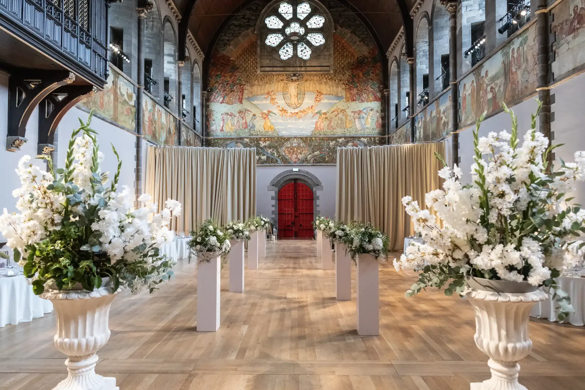 A decorated hall with a high ceiling features white floral arrangements along a wooden aisle. A red door and a mural adorn the back wall, with soft lighting illuminating the space.