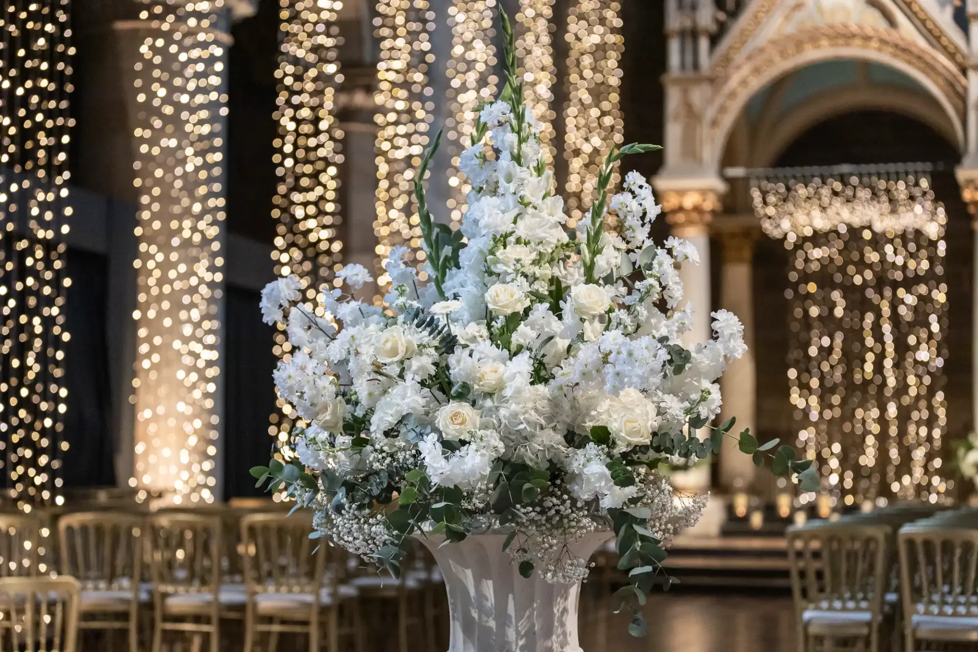 A large floral arrangement of white flowers is displayed in an ornate venue with sparkling light decorations and empty chairs in the background.