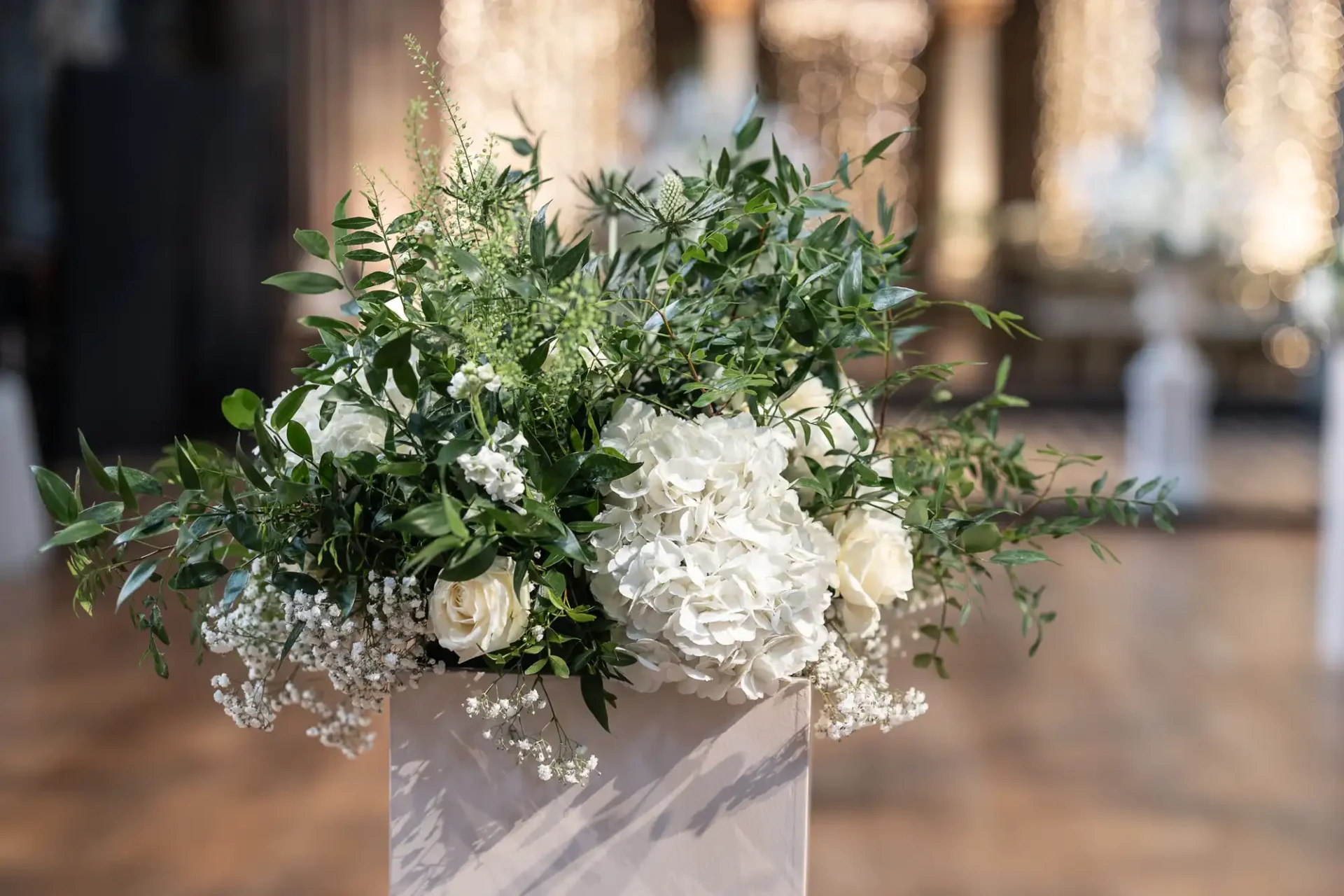 A decorative arrangement of white hydrangeas, roses, baby's breath, and greenery in a light-colored planter on a wooden floor with a blurred background.