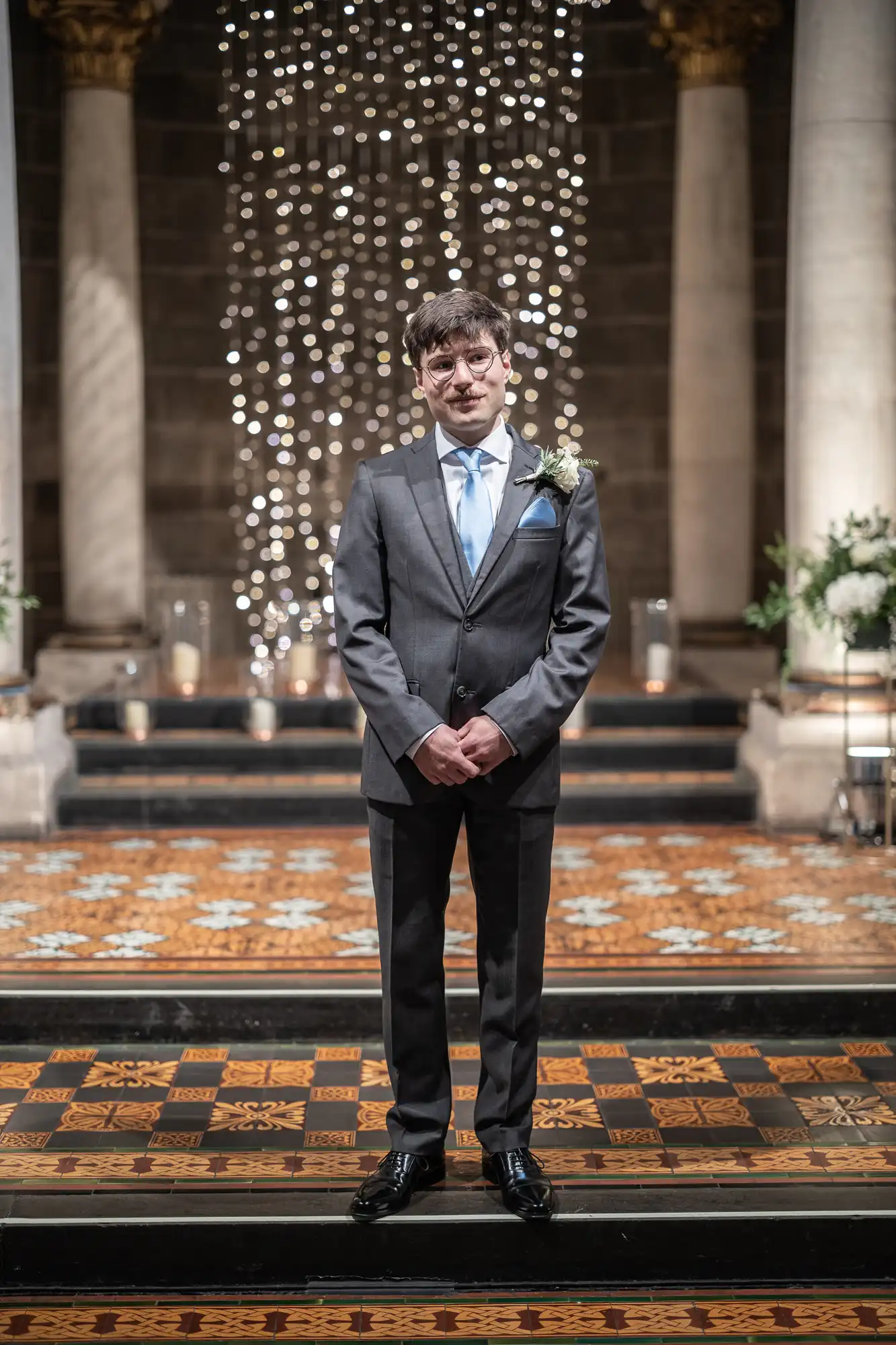 A man in a suit stands on a patterned floor in a formal setting, with columns and decorative lights in the background.