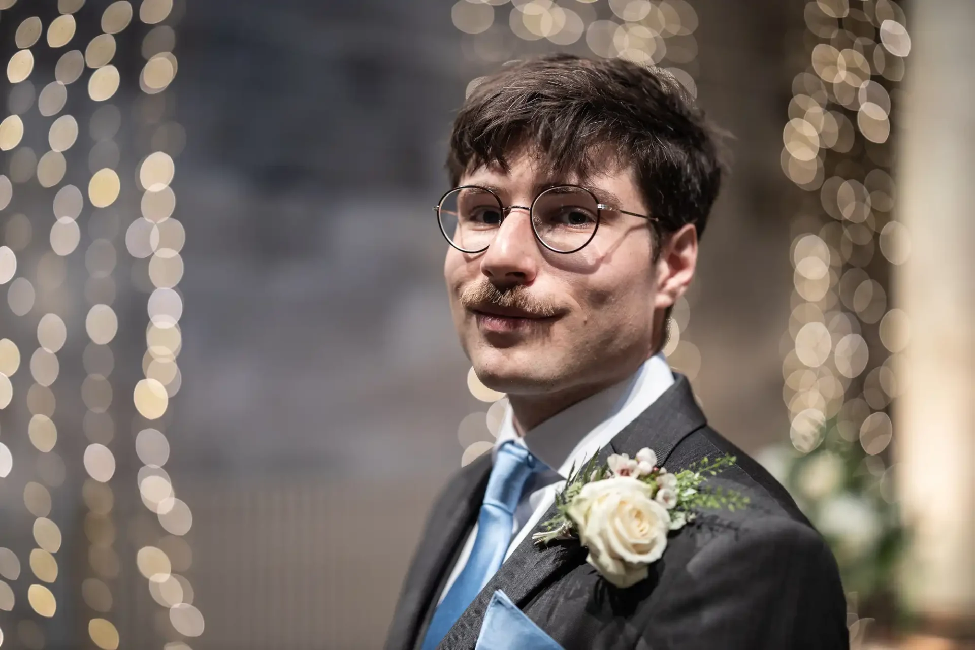 A man with glasses, a mustache, and a boutonniere on his suit jacket, stands in front of blurred lights.