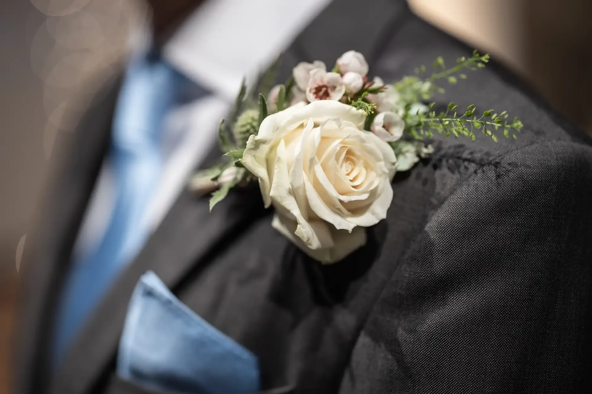 A white rose boutonniere with greenery is pinned to the lapel of a dark suit jacket, paired with a blue tie and a blue pocket square.