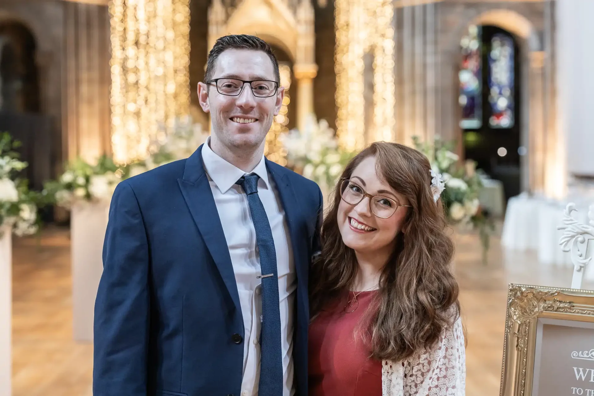 A man in a suit and tie stands beside a woman in a red dress with a white shawl. They are smiling and posing indoors with floral decorations and lights in the background.