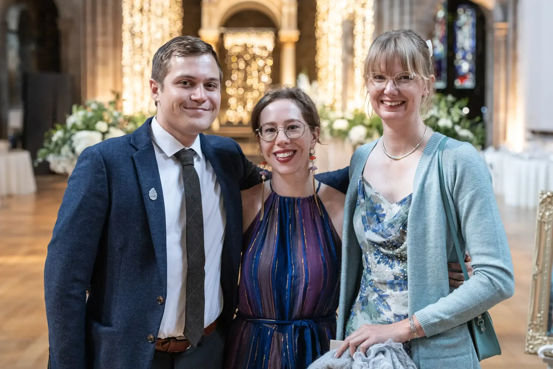 Three people pose together indoors at a decorated venue. The person on the left wears a suit, the middle person a sleeveless dress, and the person on the right a floral dress with a cardigan.