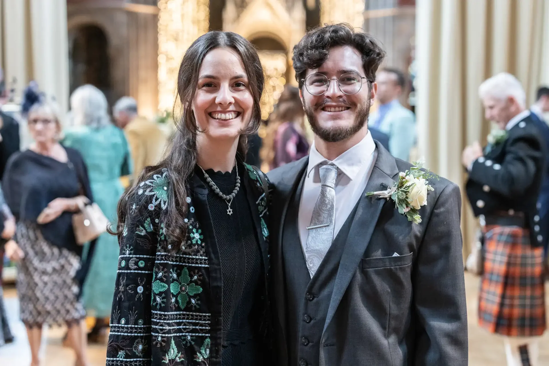 A smiling couple in formal attire pose together at an indoor event with blurred guests in the background. The man wears a suit with a floral boutonniere.