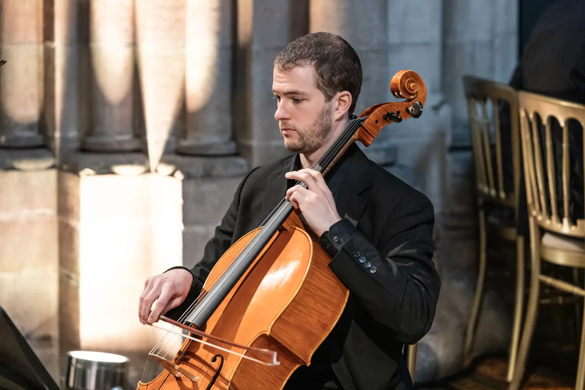 A man in a black suit plays a cello indoors, seated next to stone columns and a metal chair.