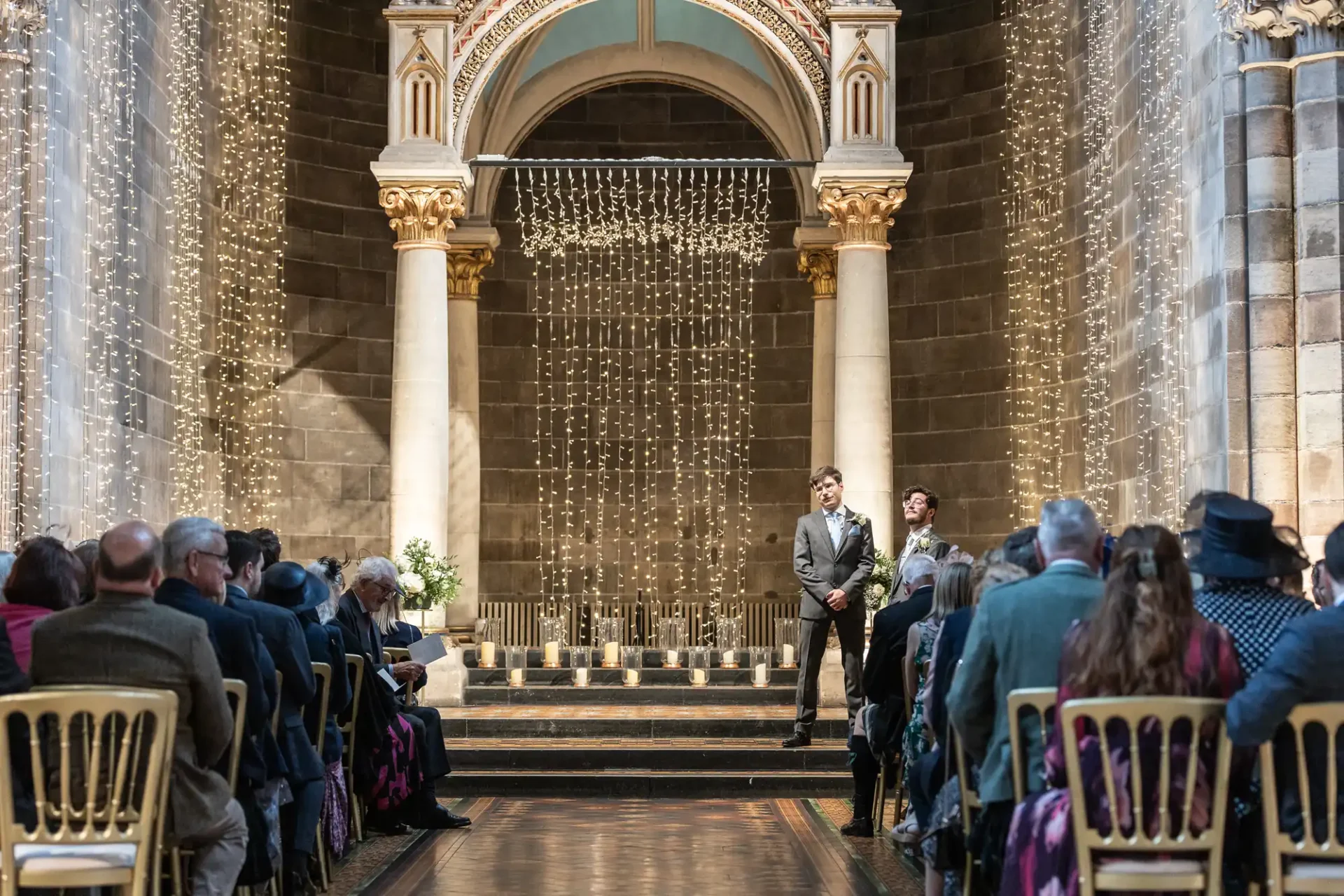 A couple stands at the altar in a decorated venue with string lights and columns. An audience sits on both sides, watching the ceremony.