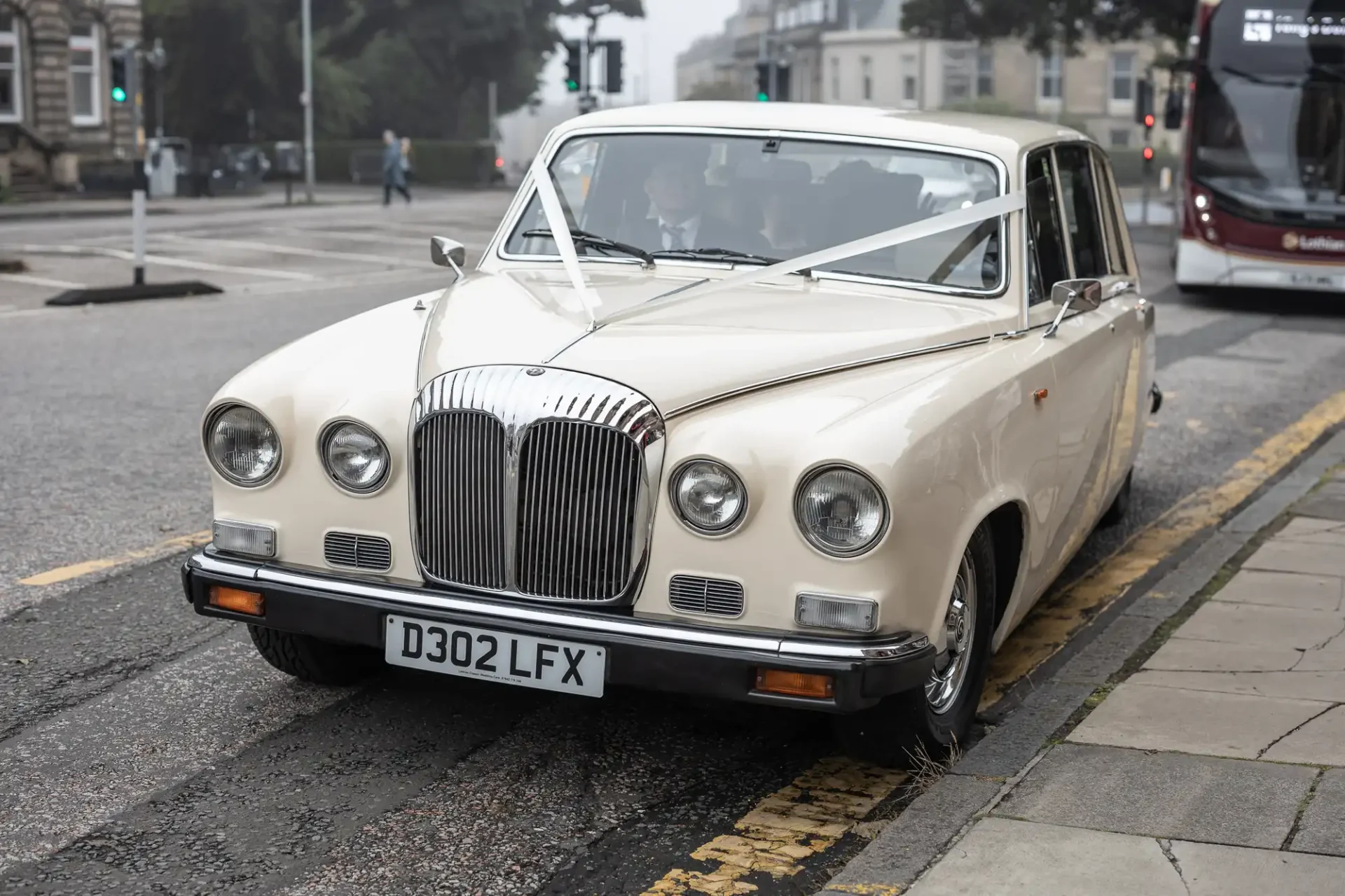 A vintage white car with a decorative ribbon is parked on a street near a bus stop.