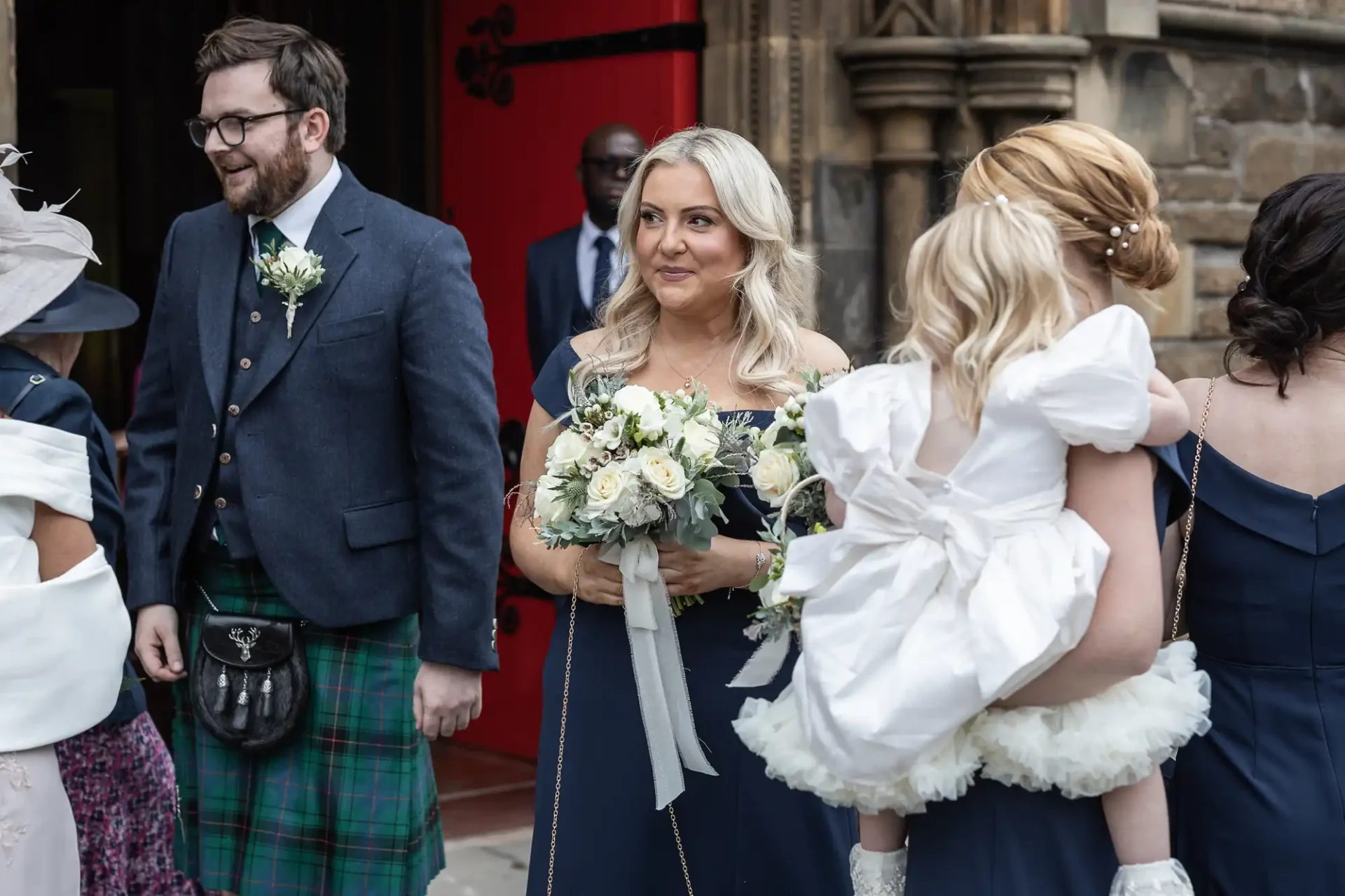 A group of people in formal attire stand outside a building. One woman holds a bouquet, and another carries a child wearing a white dress. A man in a kilt is also present.