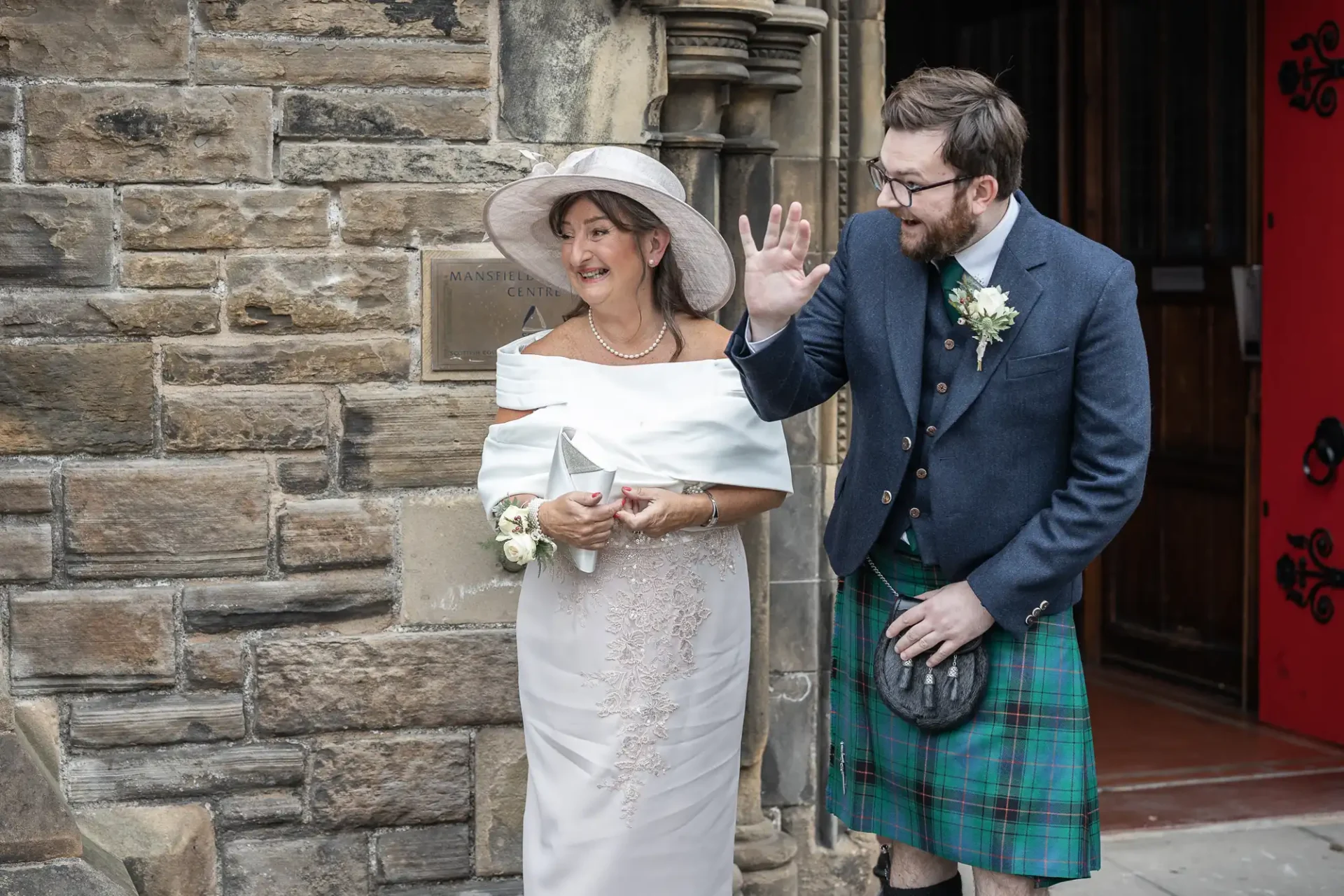 A woman in a white dress and hat stands next to a man in a kilt and jacket, who is waving. They are in front of a stone building with a red door.