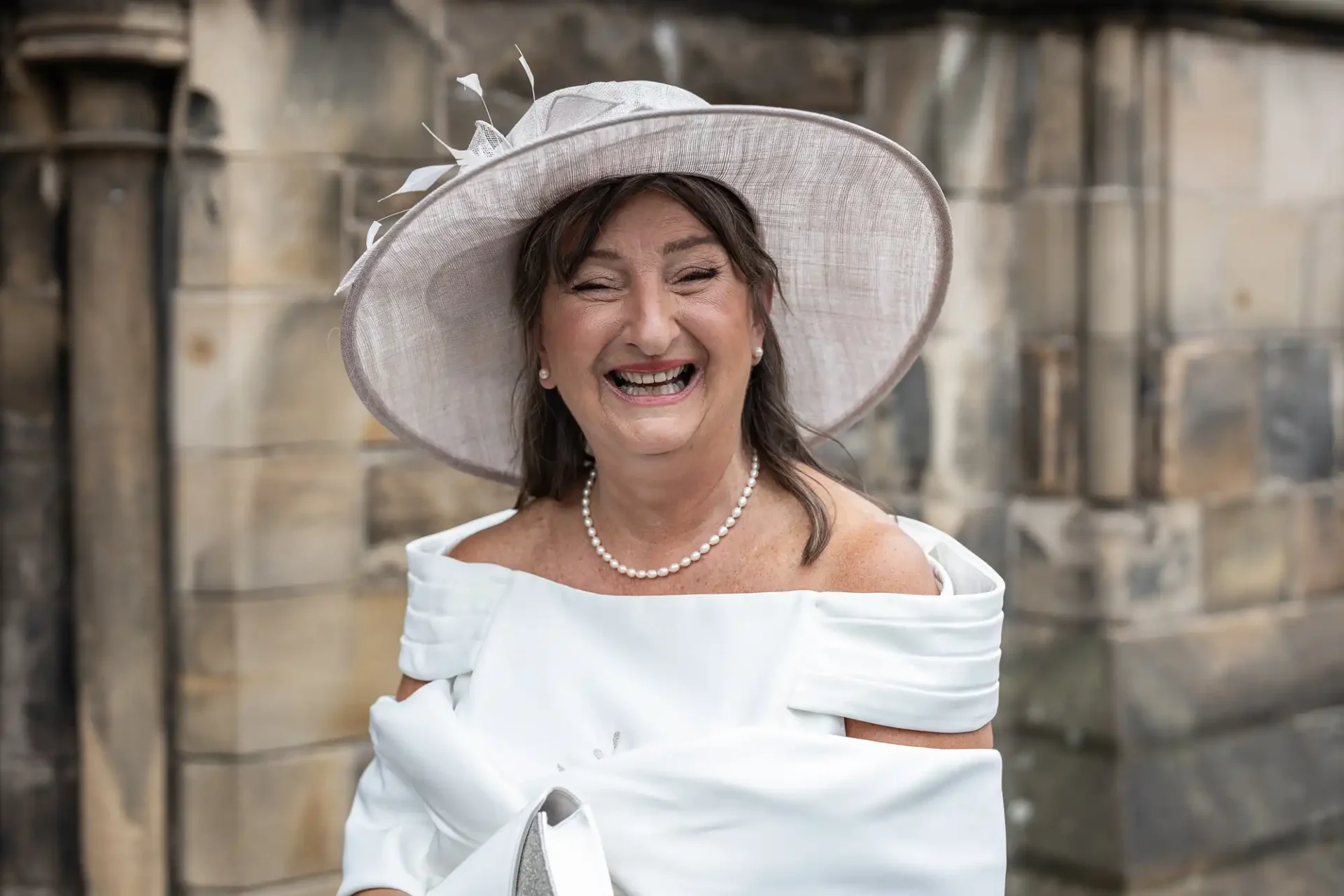 Smiling woman in a white dress and stylish hat standing outdoors against a stone wall.