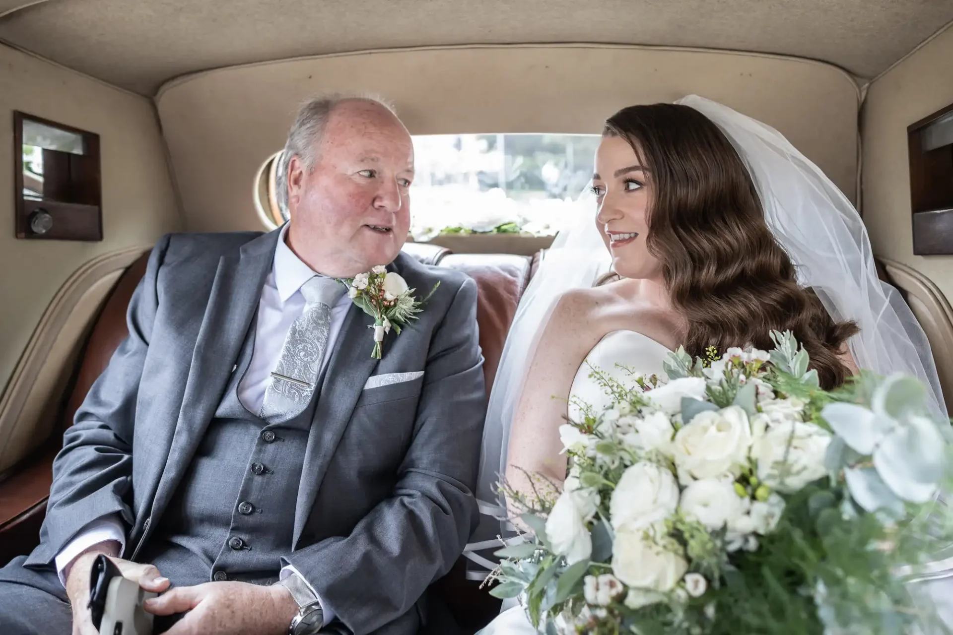 A bride and an older man sit together in a car. The bride holds a bouquet and wears a veil. Both are dressed formally and seem to be engaged in conversation.