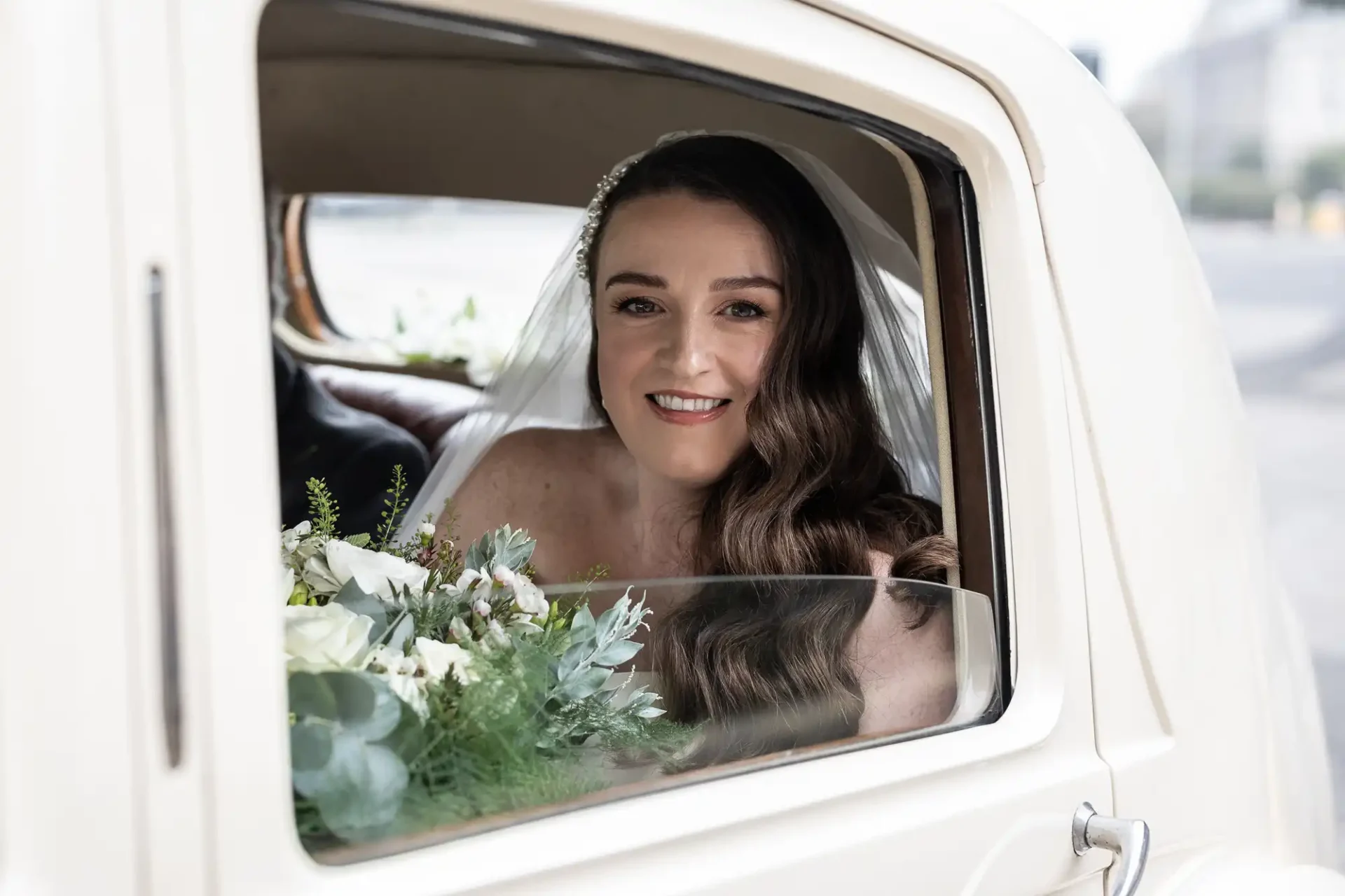 Bride smiling, sitting in a vintage car, looking out the window. She's holding a bouquet with white flowers and greenery, wearing a veil and white dress.