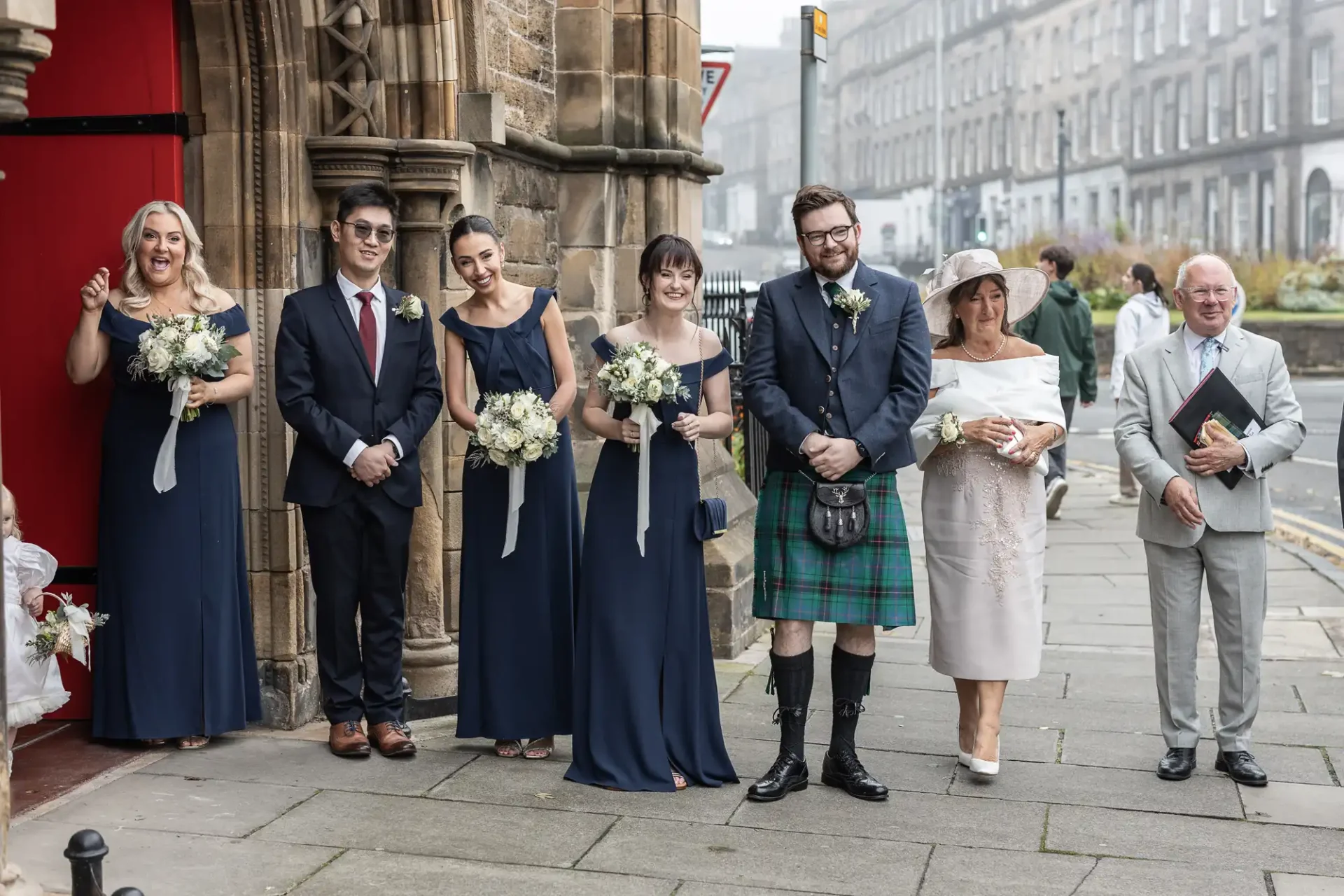 A group of people stand outside a stone building. Four women are holding bouquets, and one man is wearing a kilt. They are dressed in formal attire.