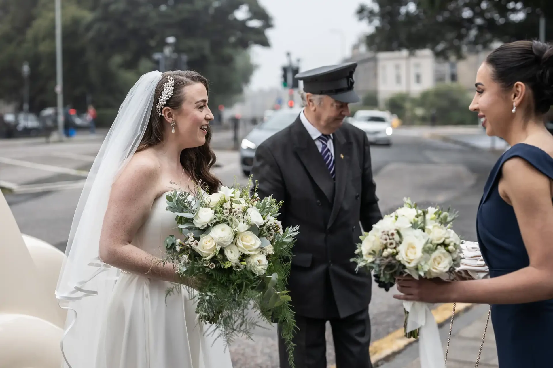 Bride in white gown holding a bouquet, standing with a bridesmaid in a dark dress and a chauffeur in a uniform, near a car on a cloudy day.