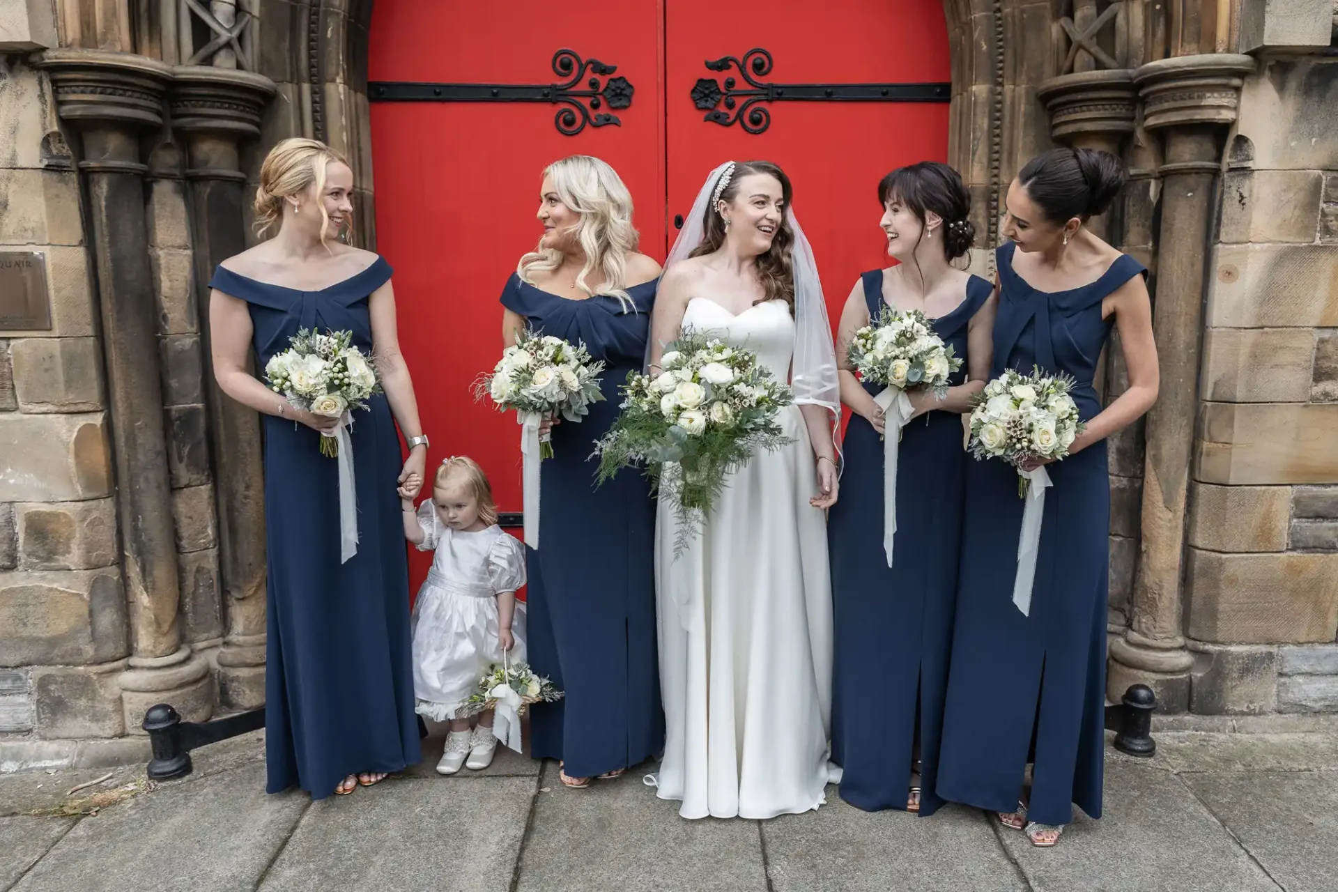 Bride and bridesmaids in navy blue dresses stand smiling in front of ornate red doors, holding bouquets. A young girl in a white dress stands among them.