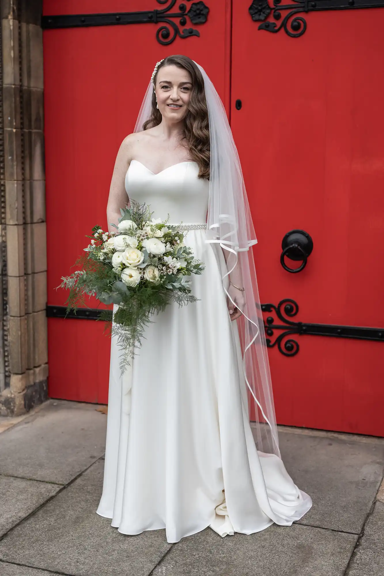 Bride in a white dress and veil holds a bouquet of white flowers and greenery, standing in front of a large red door with black accents.