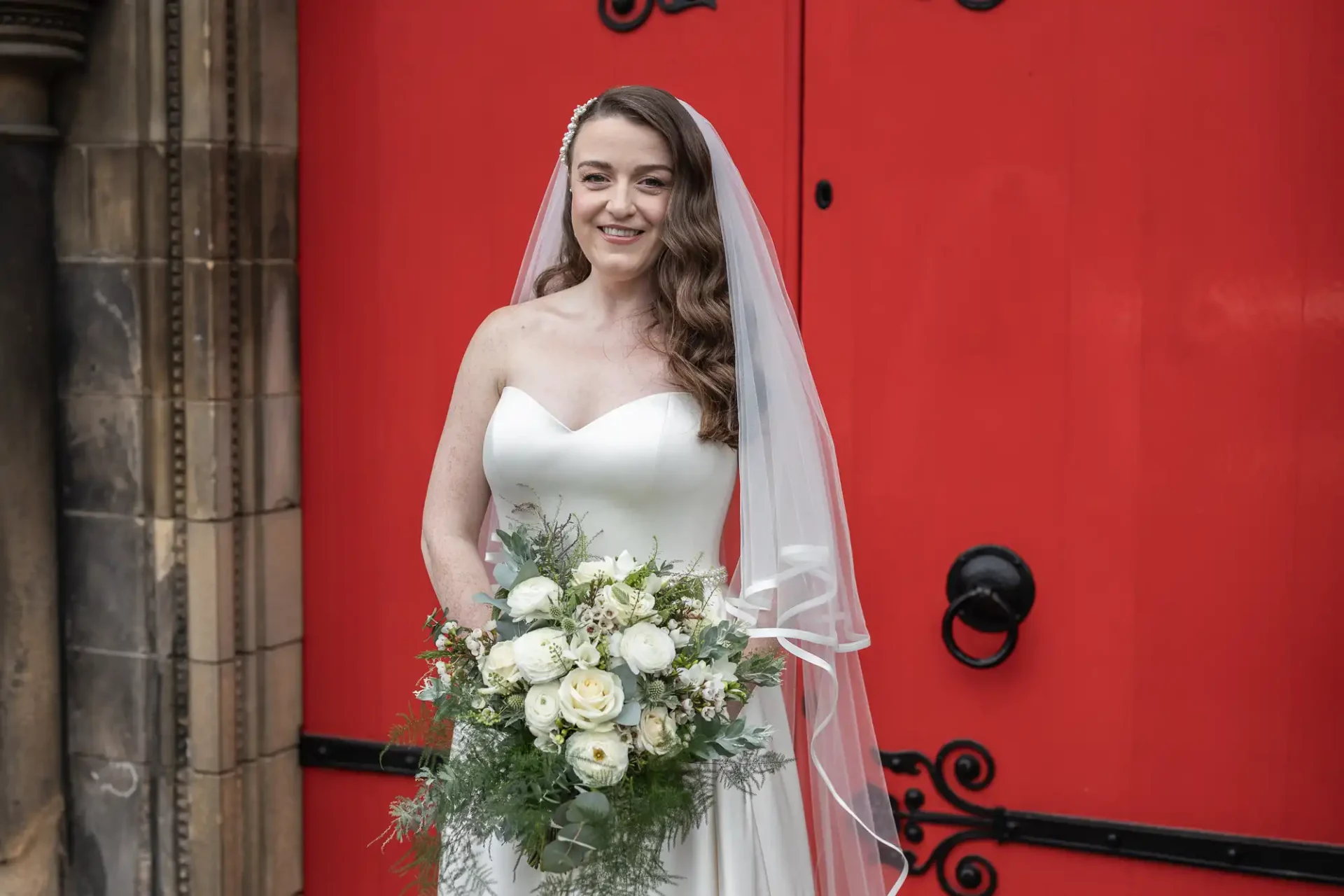 Bride in a white dress and veil holding a bouquet of white flowers, standing in front of a large red door.