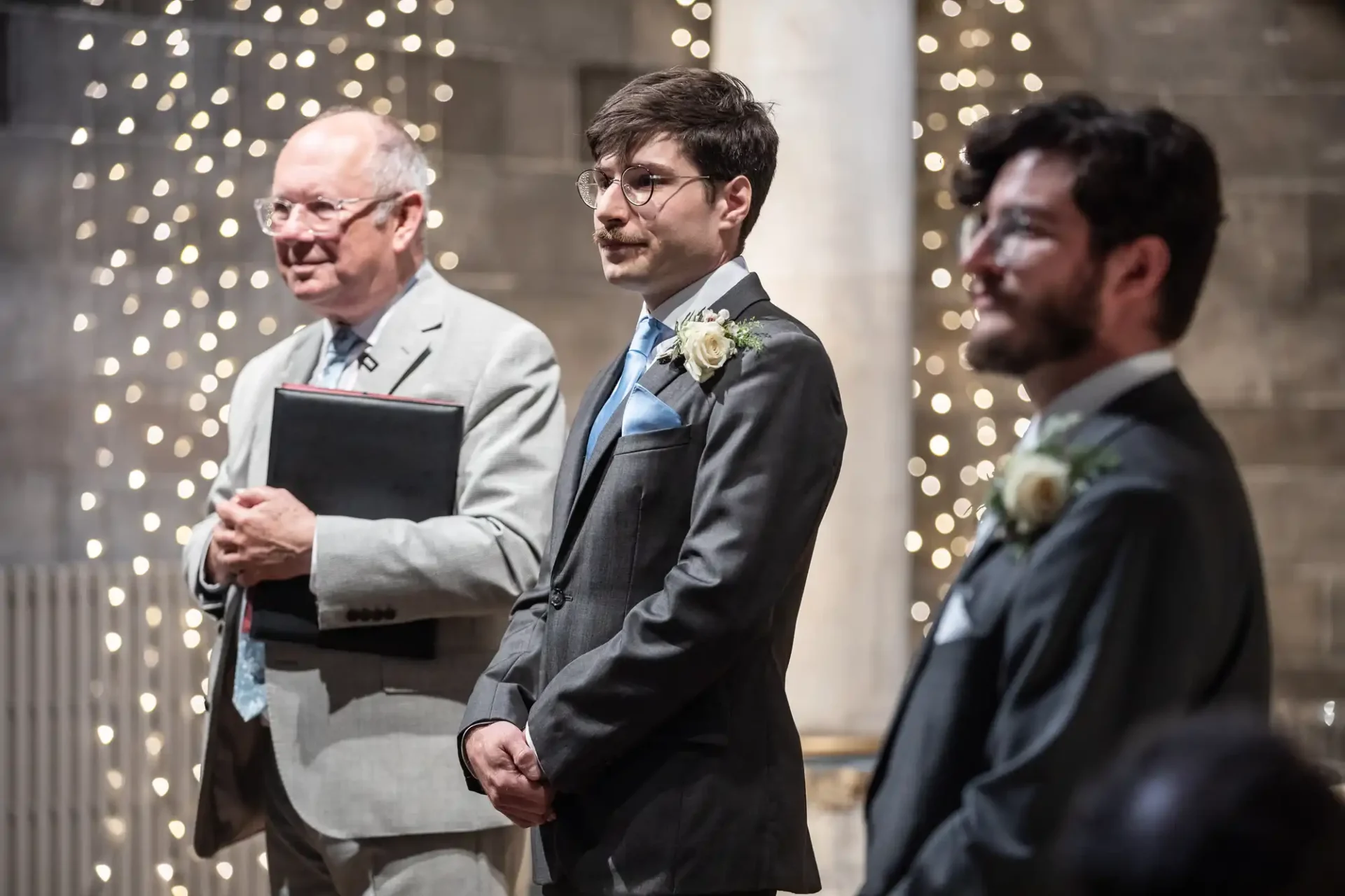 Three men, two in suits with boutonnieres and one holding a folder, stand indoors against a backdrop of decorative lights.