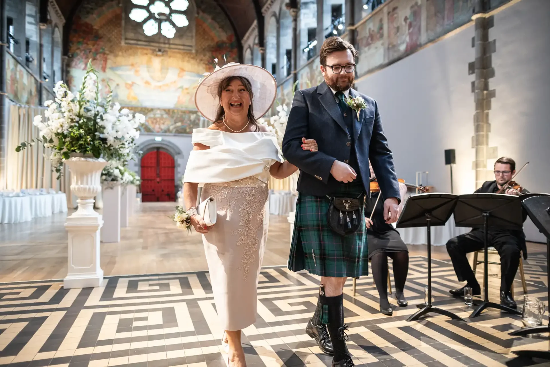 A woman in a light outfit and hat walks arm-in-arm with a man in a kilt down an aisle, with musicians playing string instruments in the background.