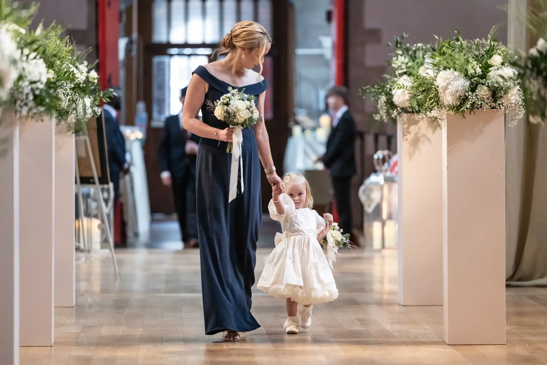 An adult in a blue dress holds hands with a child in a white dress as they walk down an aisle decorated with flowers.